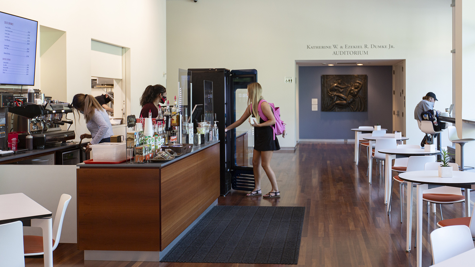 view of the museum cafe with a customer at the counter reching to hand payment to the barista behind the counter. There is another customer sitting at the long bar in the wondow.