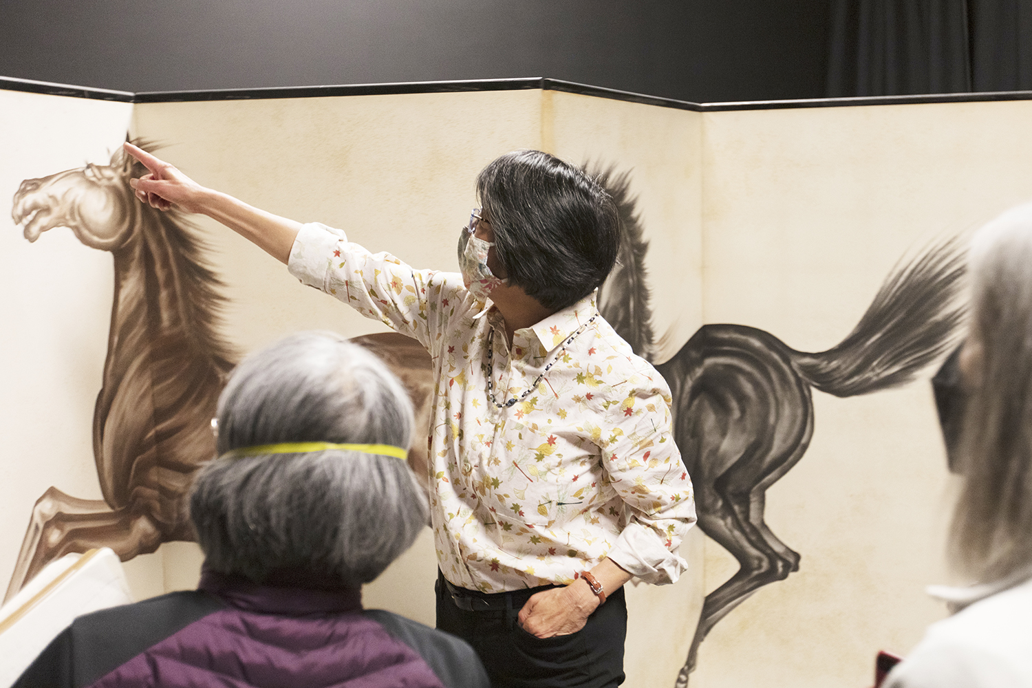 A tall Japanese style screen made of four panels with two large horses, one black one brown painted in large, gestural strokes arcoss the panels. Kimi Hill stands in from on the screen gesturing to a detail for the crowd of people looking on.