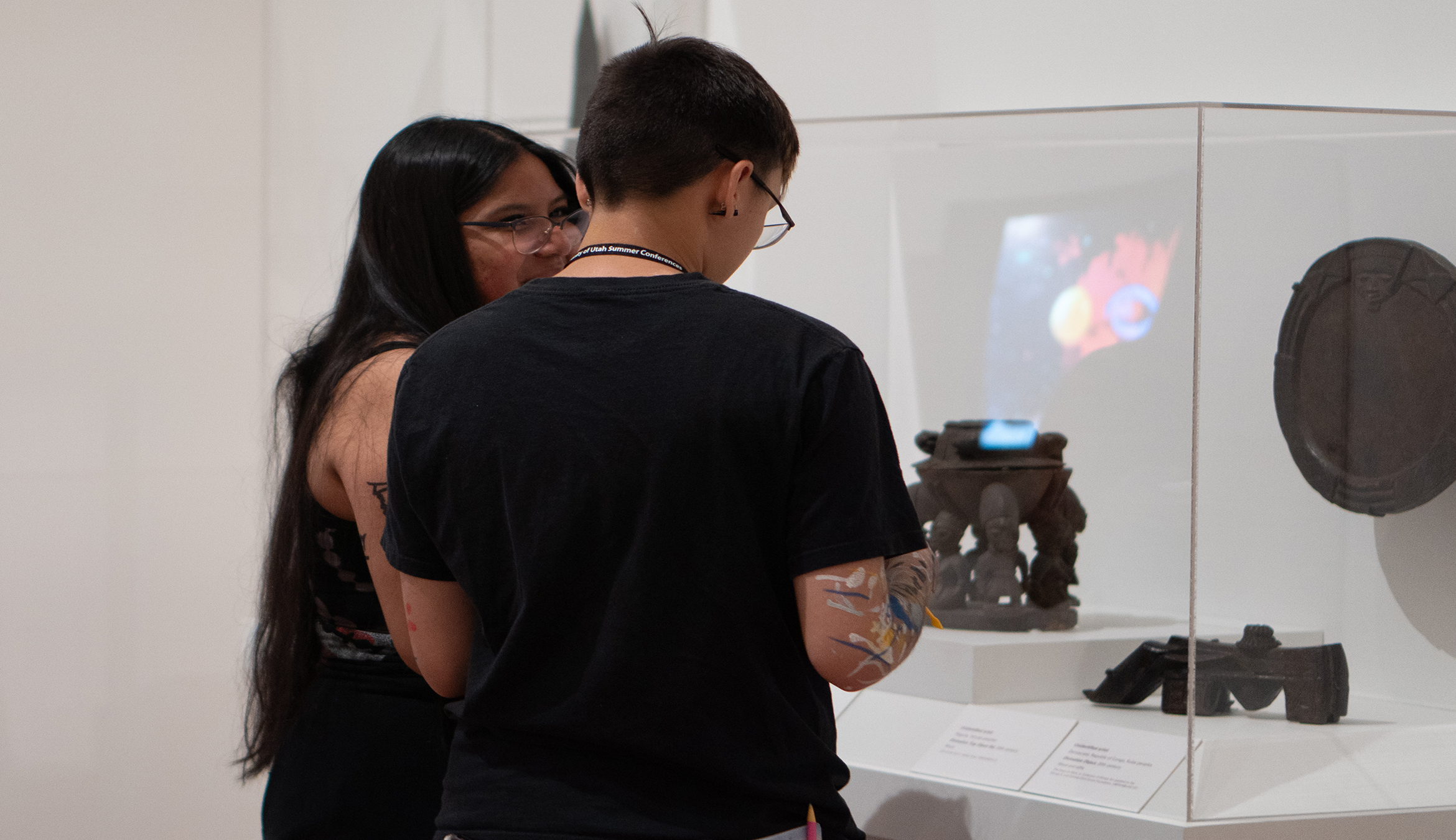 Two young people with dark hair talk and look at artifacts in a display case.