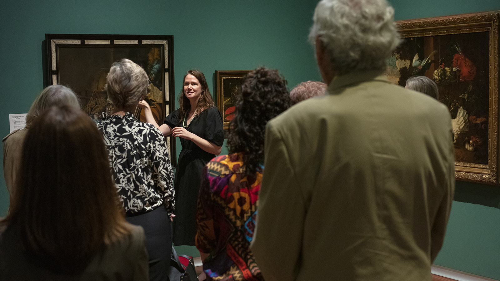 A group of museum visitors gathered around a female curator as she gesture towards and oil painting the European gallery