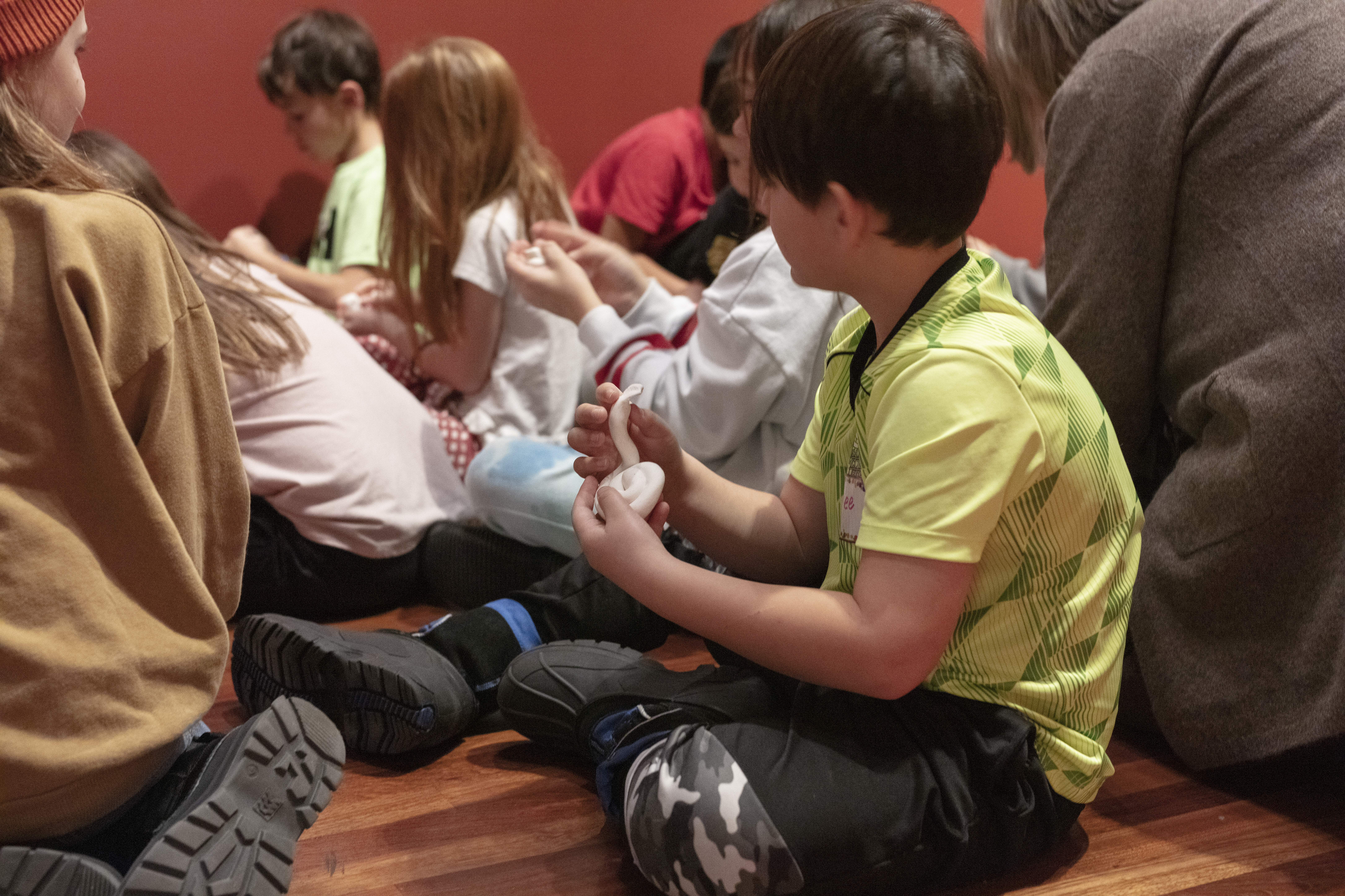 A group of children sit on the floor sculpting clay. A boy with dark hair in a green shirt holds white clay shaped like a coiled-up snake
