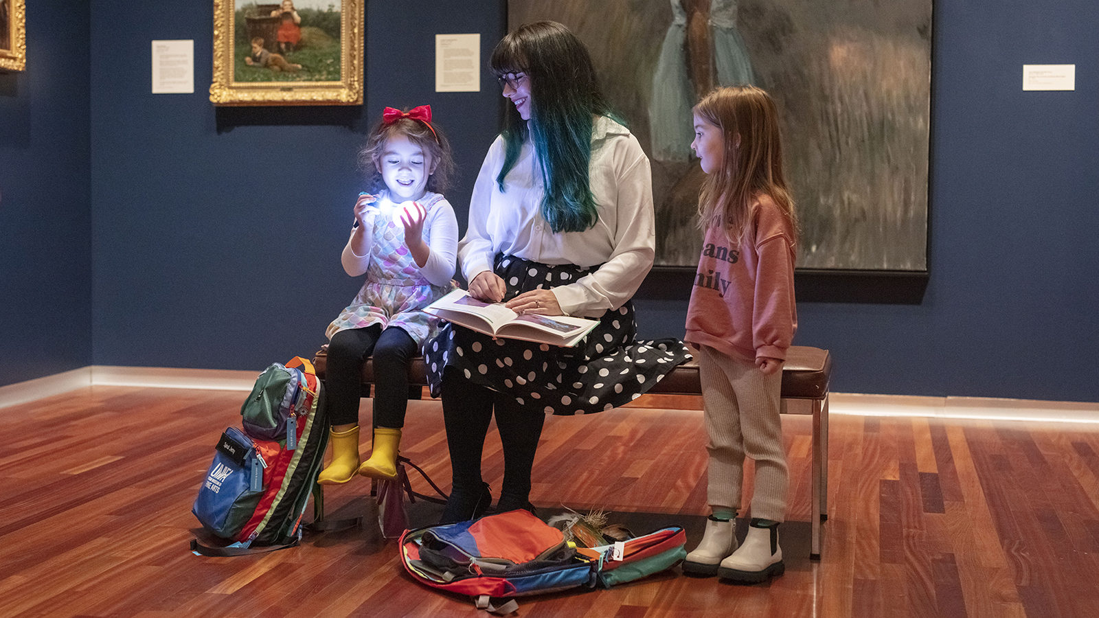 a mother sits on a bench in the UMFA European gallery flanked by two young girls. One girl in is shining a flashlight on her had and the other looks at her laughing. there are two UMFA backpacks at their feet.