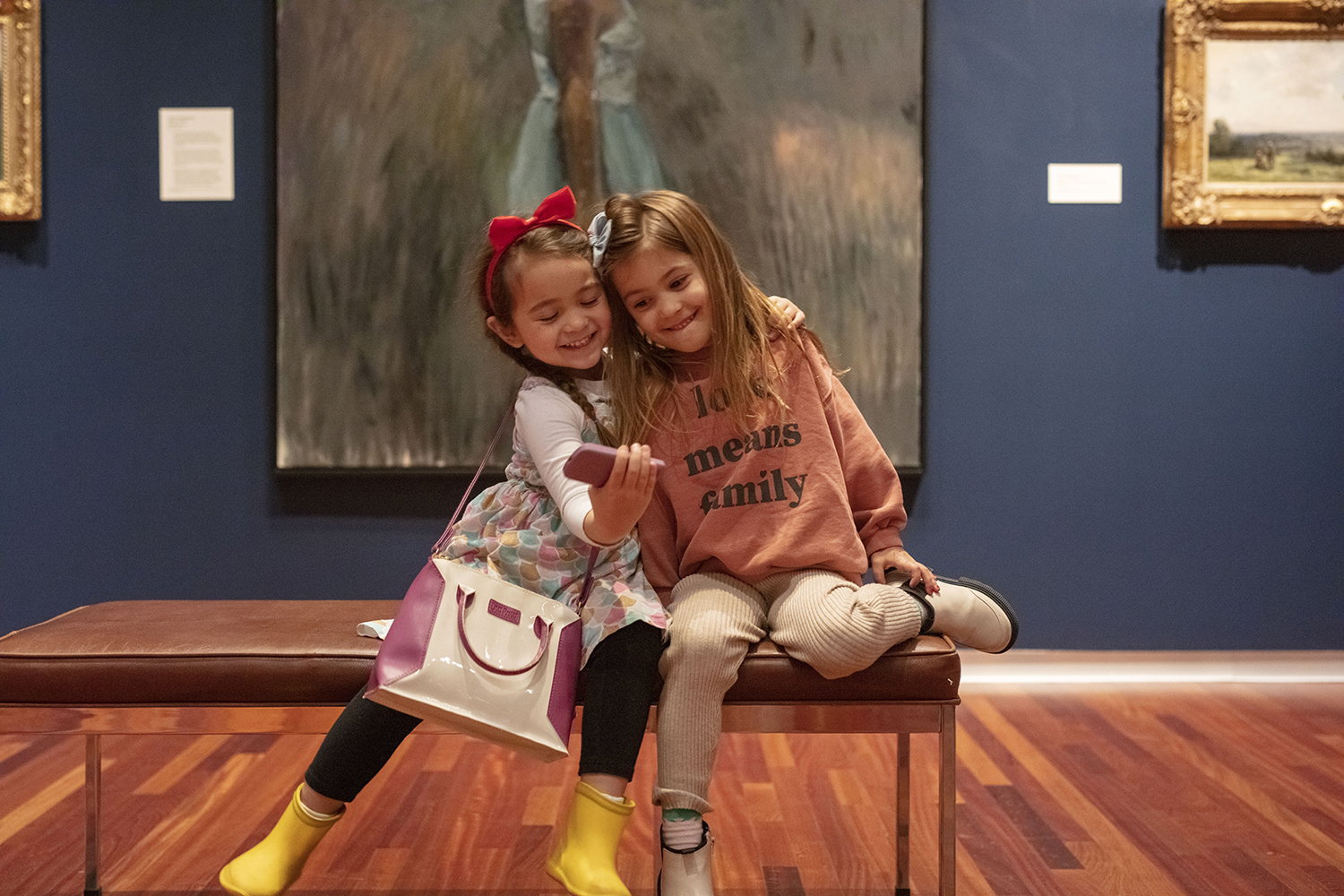 two little girls sit on a bench in a museum gallery pretending to take a selfie with a toy phone