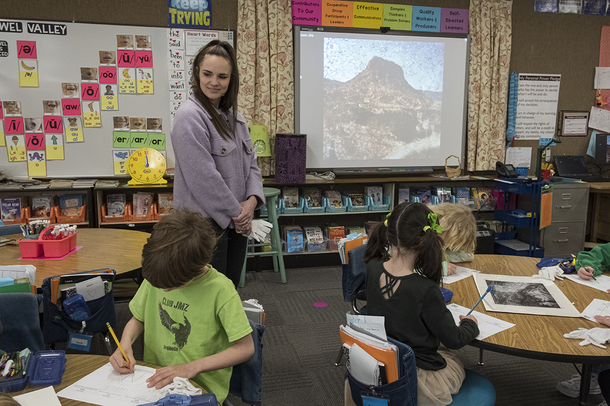 Katie Seastrand stands in a classroom with first graders sitting at round tables there is a screen with a projection of a landscaped photograph of a red rock hill in the back ground and photos on the tables with the children