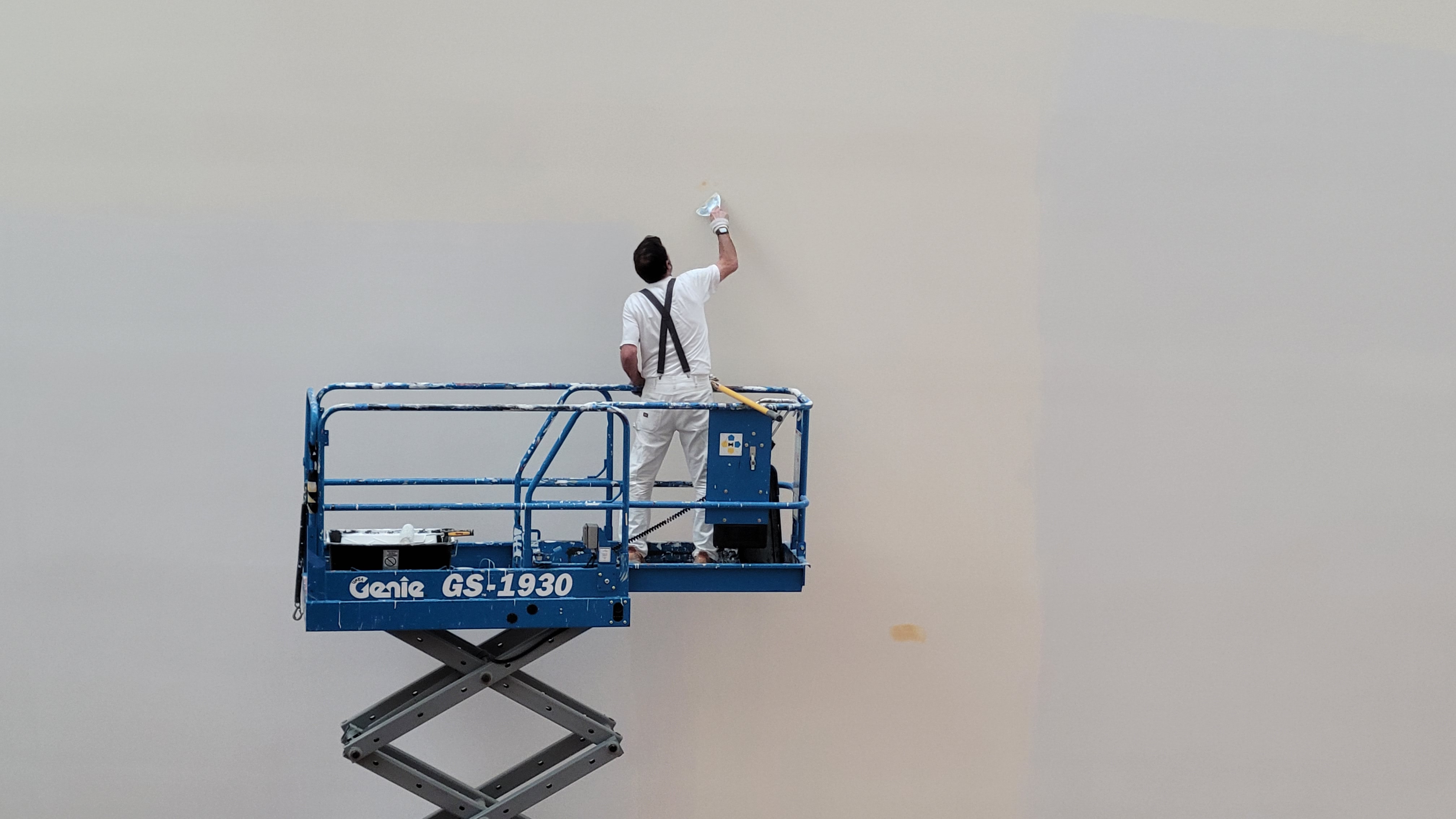 A person stands on a "Genie" brand lift and repaints a wall.