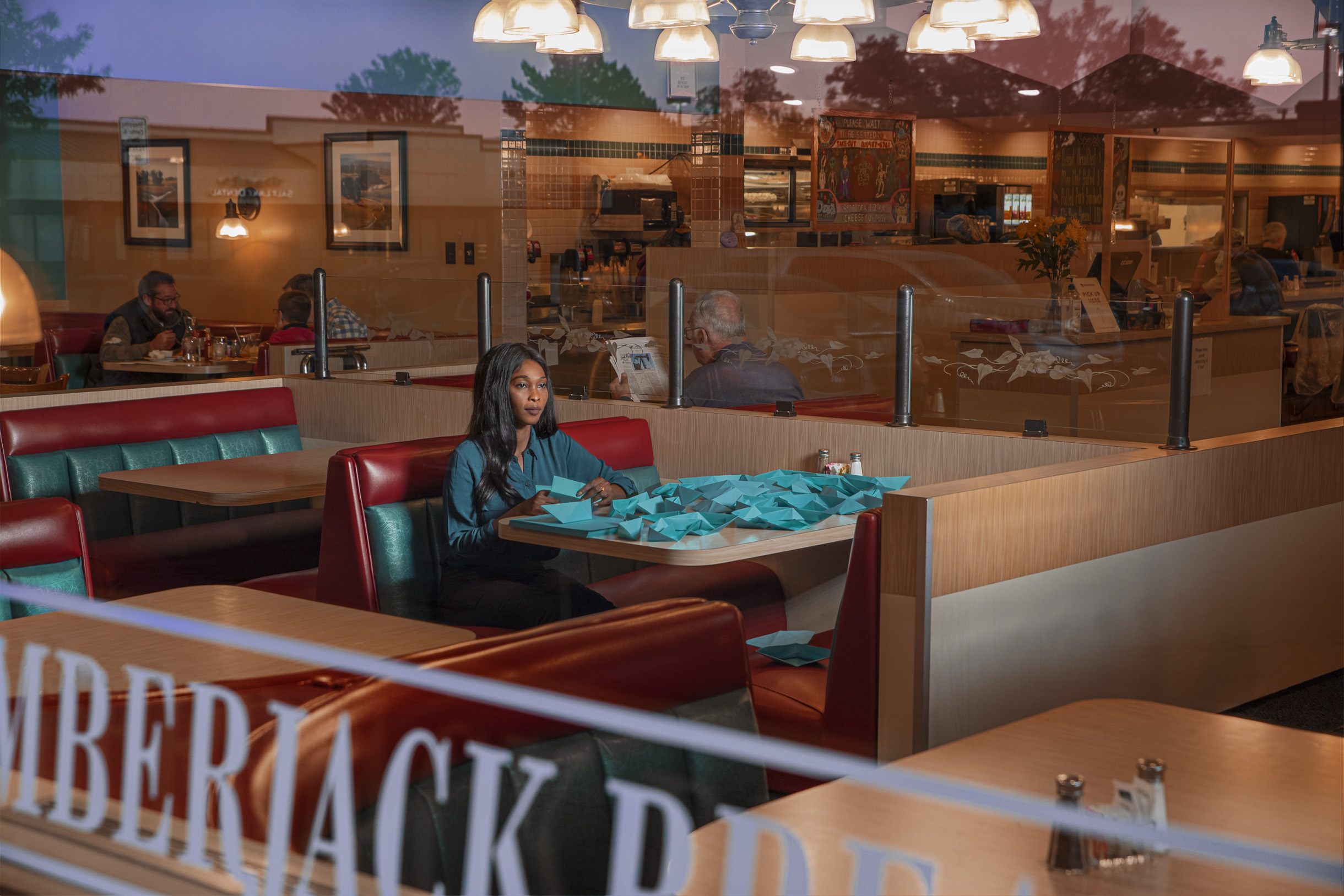 a woman sits in a diner on a blue and red booth seat. She is folding blue paper and the table is covered in folded blue paper. The photograph is taken from outside looking in through the window.   