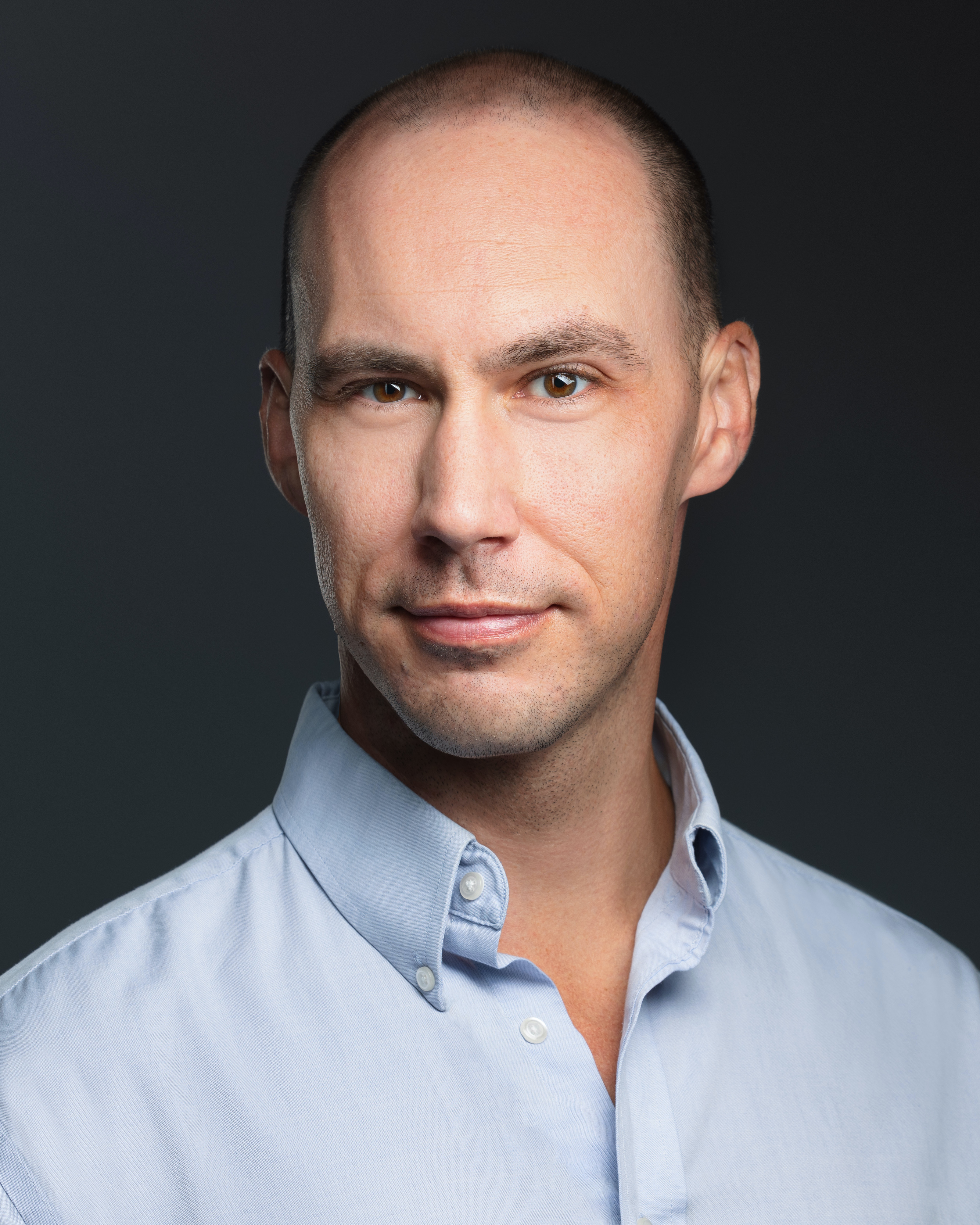 A headshot photo of a man with a shaved head, wearing a light blue button up shirt.