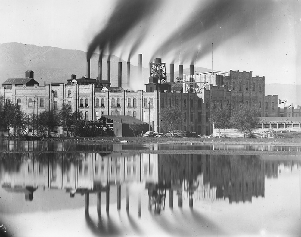 vintage black and whit images of a factory or industrial building with 8 smoke stacks emitting smoke, the building is reflected in a body of water before it