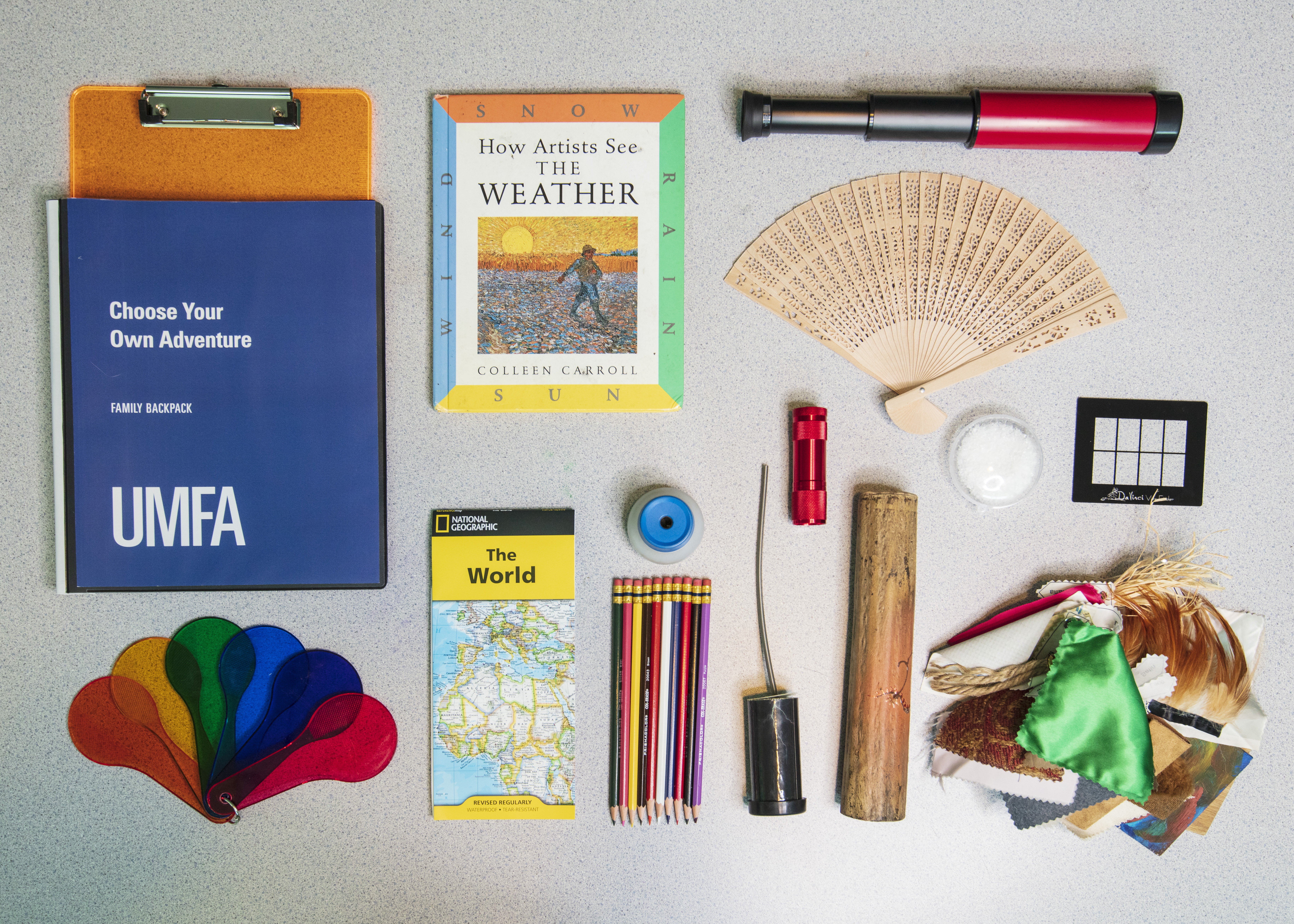 The contents of a UMFA sensory backpack laid out. There is a map, a telescope, color swatches, a bundle of different fabrics and textures, pencils, and more.