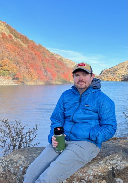 A white man sitting outside on a rock wearing a blue puffer jacket.