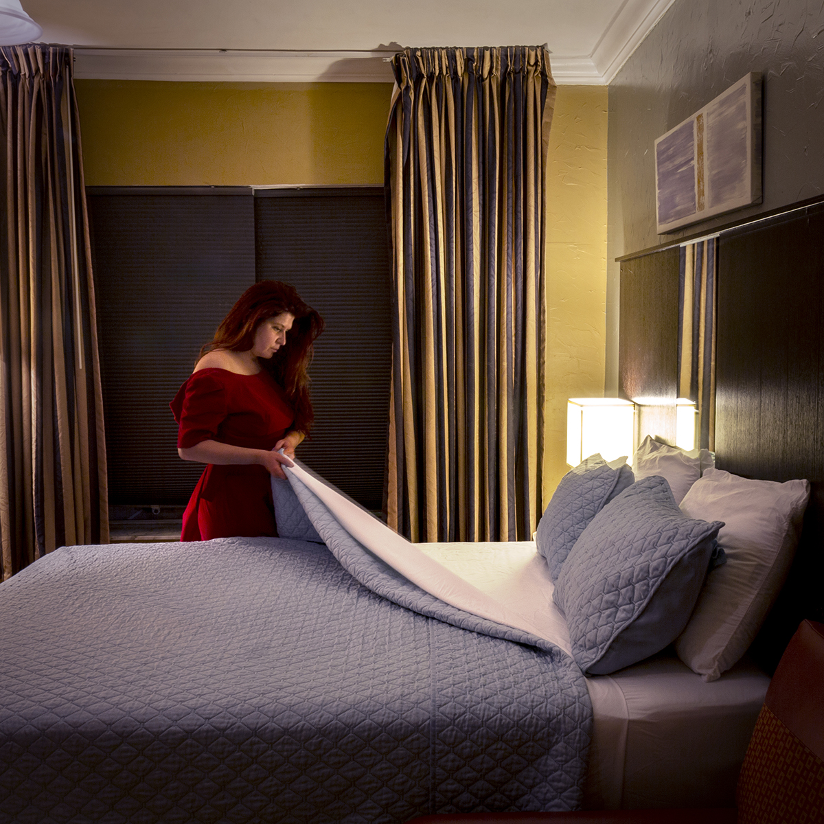 a woman in a red dress stands in a hotel room. She is pulling back the blue covers on the bed. The blinds on the window behind her are closed and a lamp on the bedside table is turned on.  