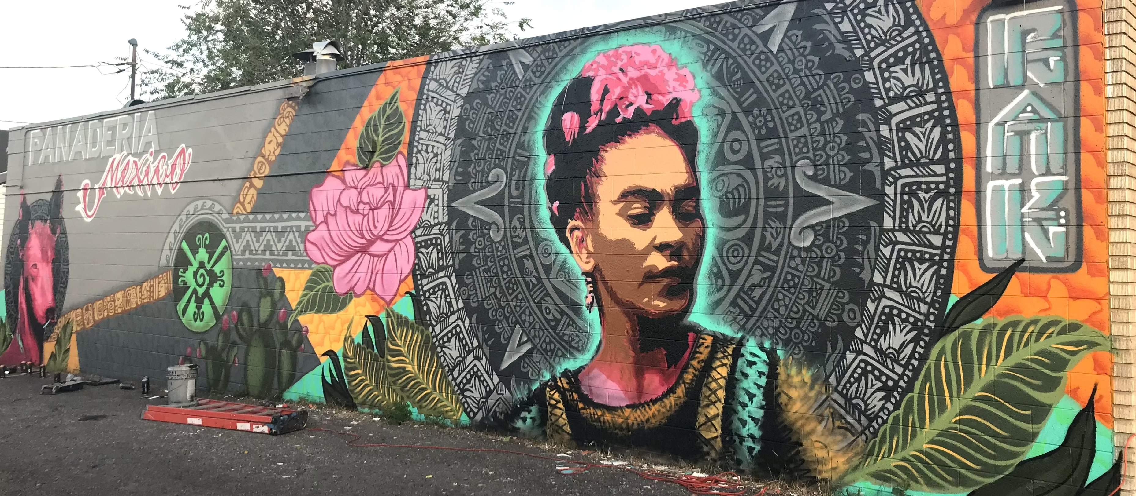 A mural of Frida Kahlo enciricle by an Aztec-looking mandala with the words Panaderia Mexico on the left side of the mural on a cinderblock wall 