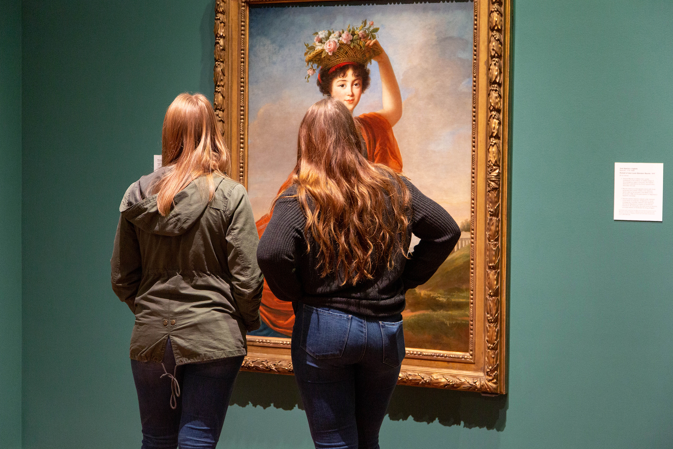 Two women with long hair stand and stare at a large, gold-framed oil painting of a woman with a basket of flowers on her head.