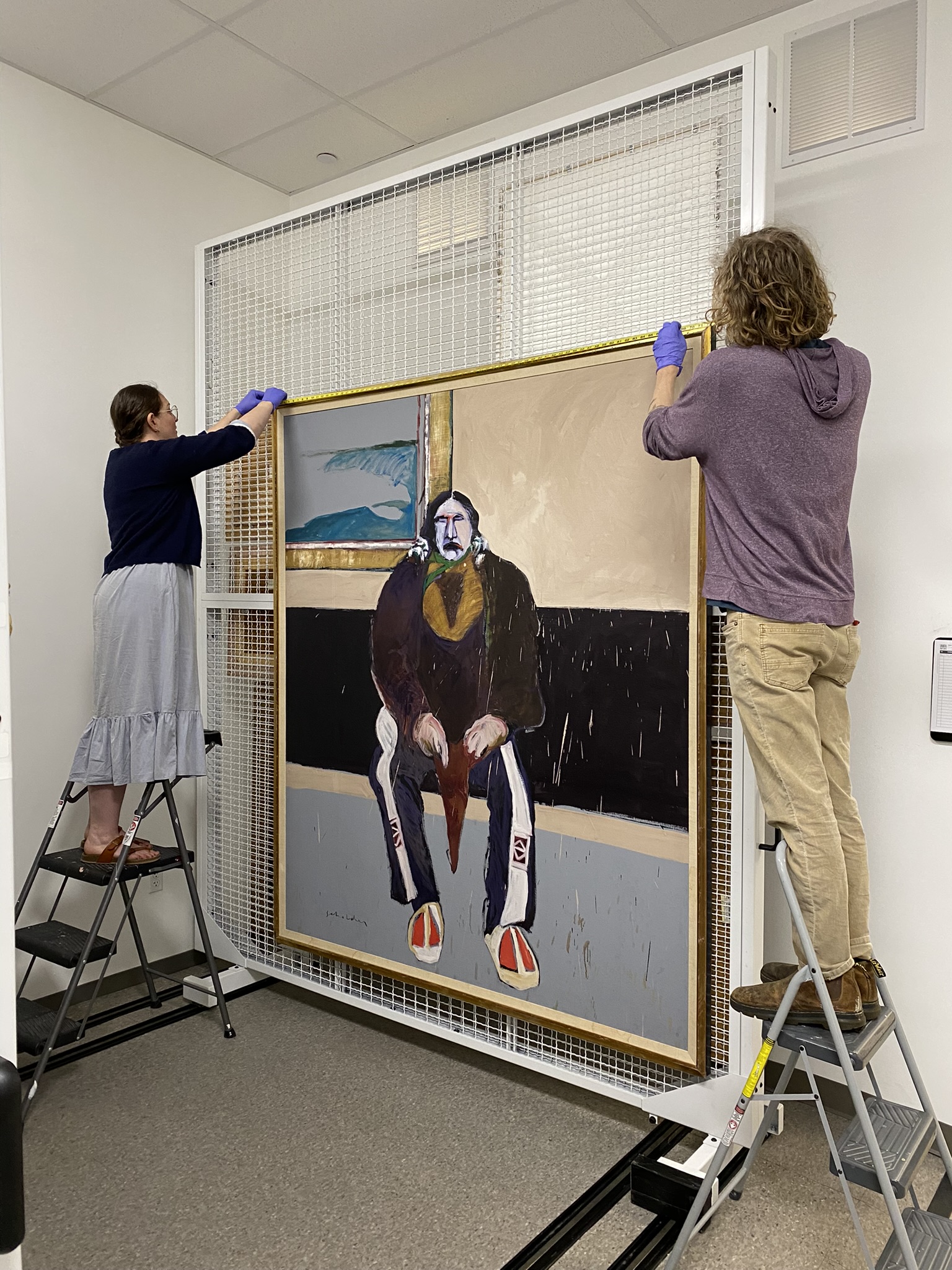 Two people stand on step stools on either side of a painting and secure it for storage.