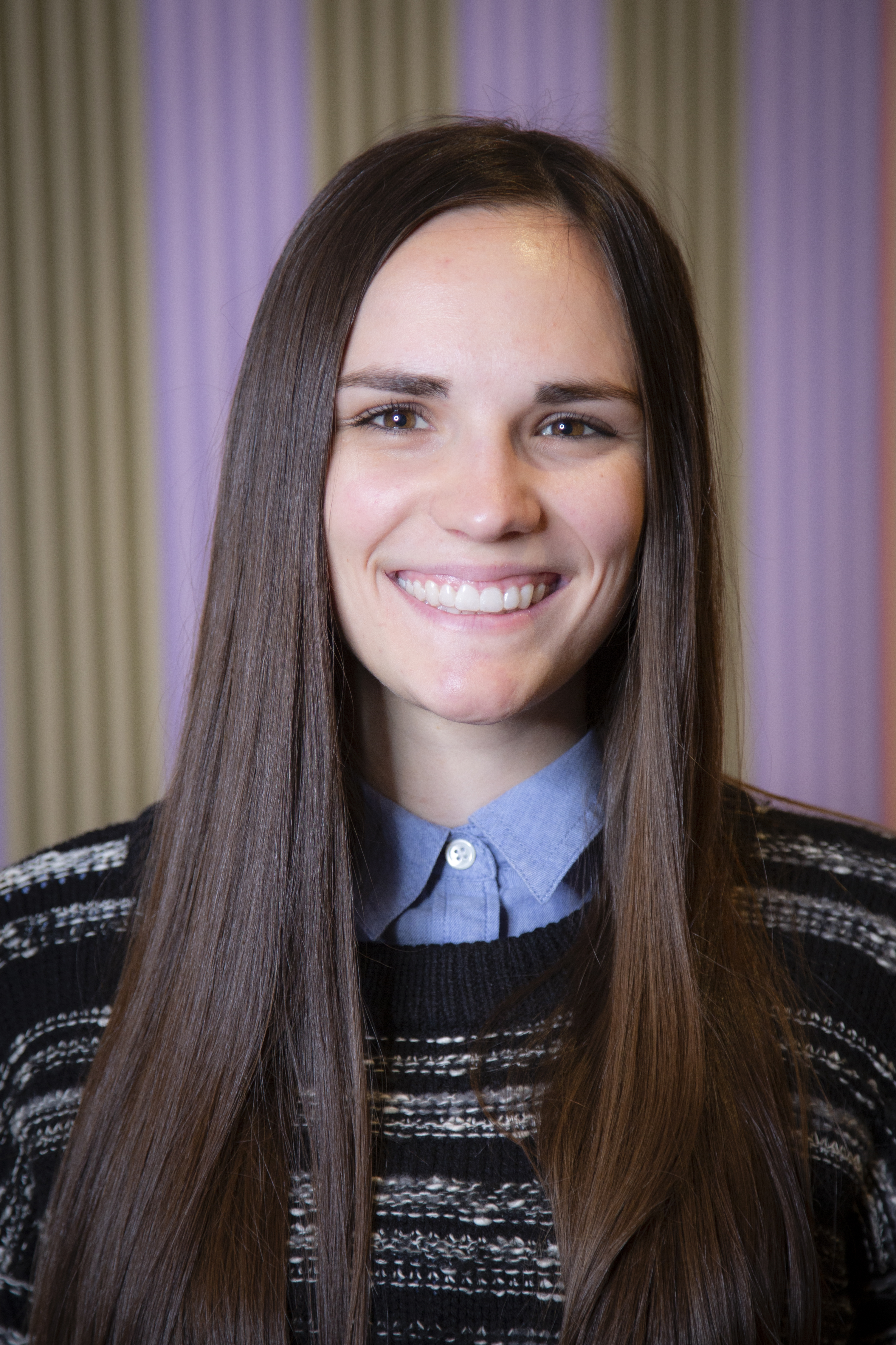 a head shot of Katie Seastrand looking directly at the camera smiling she has long dark brown hair and is in front of a painting of various colored vertical stripes. 