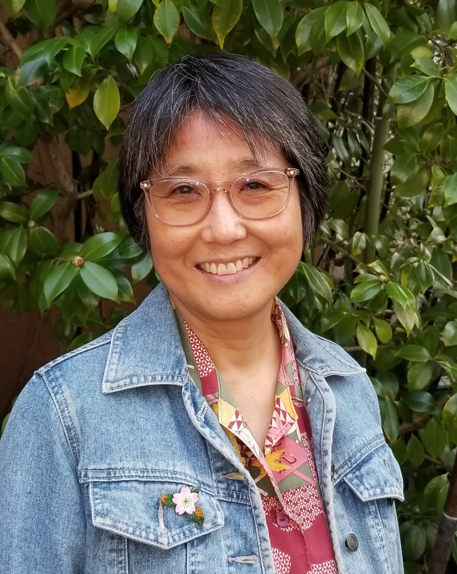 A photo of Kimi Hill smiling. She is wearing glasses and a denim jacket.