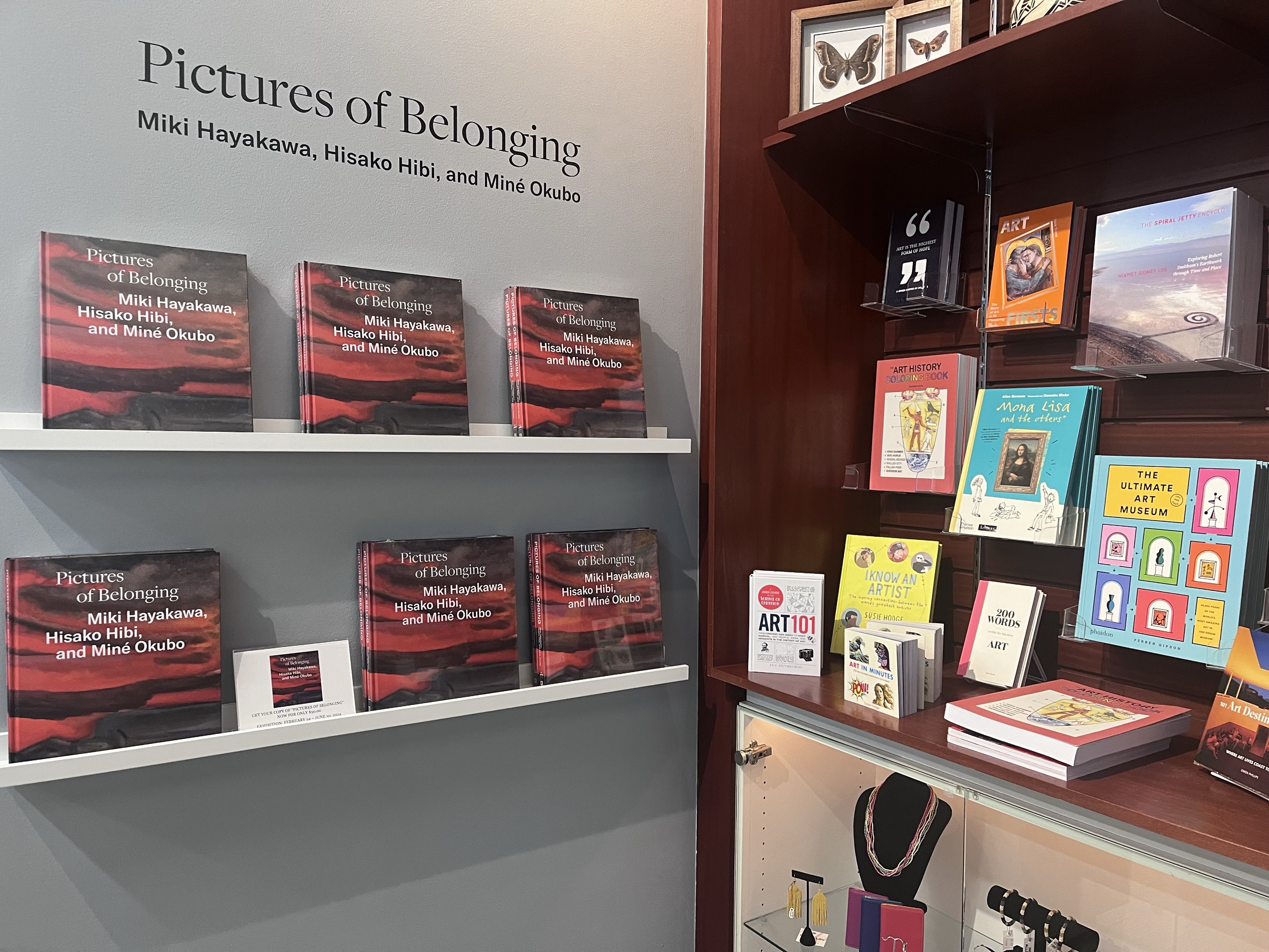 A group of the same book with a black and red cover sit on shelves on a light grey wall beneath a sign that says Pictures of Belonging. There are more books and miscellaneous items on a shelf to the left.