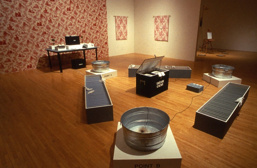 A room with a red toile adorning the walls. A table, basins, and various other objects are arranged on the floor.