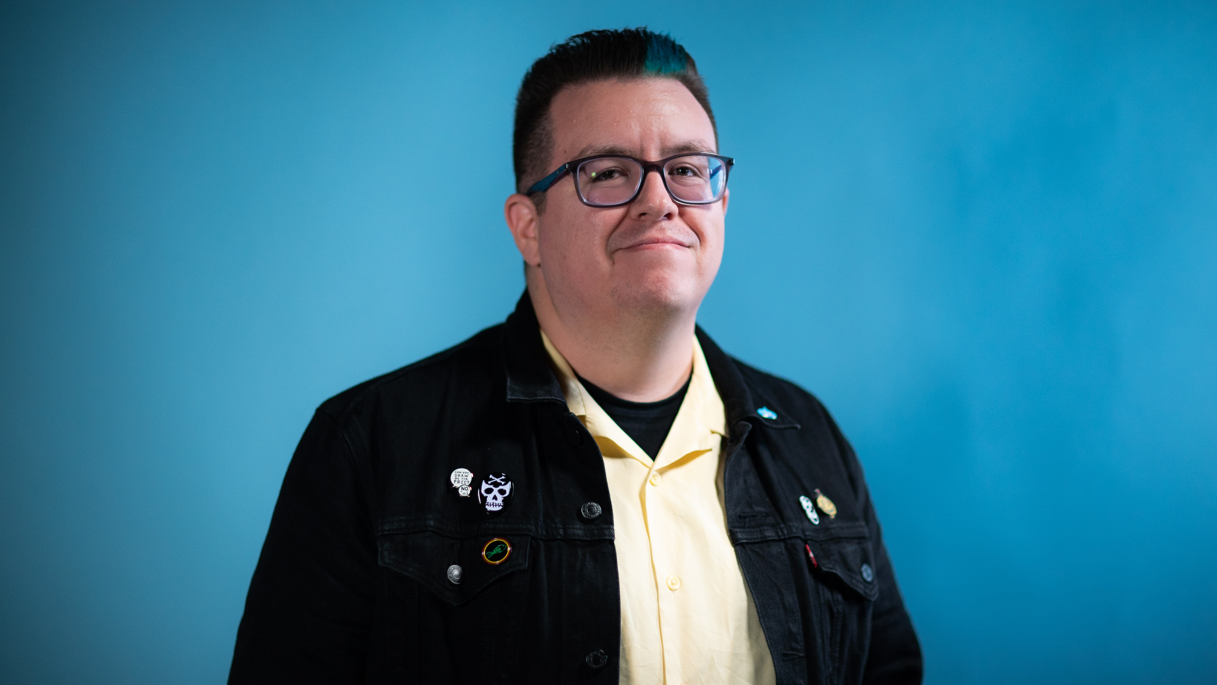 A photograph of artist Ricky Vigil on a blue background. He is wearing a white collared shirt and a black jacket with pins on it.