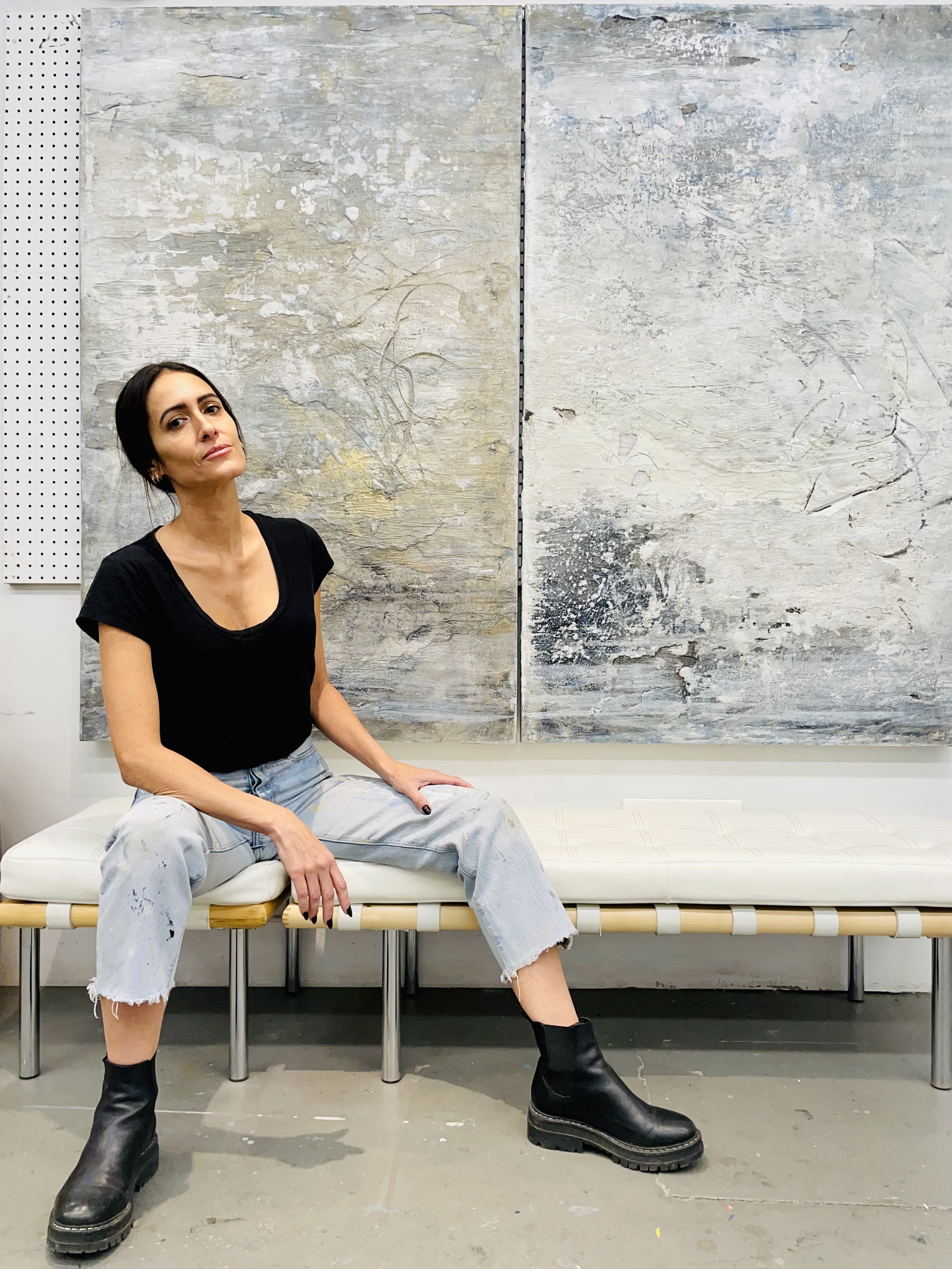 A photograph of artist Samantha da Silva seated in front of a painting. She is wearing a black shirt and black boots with cropped light wash jeans.