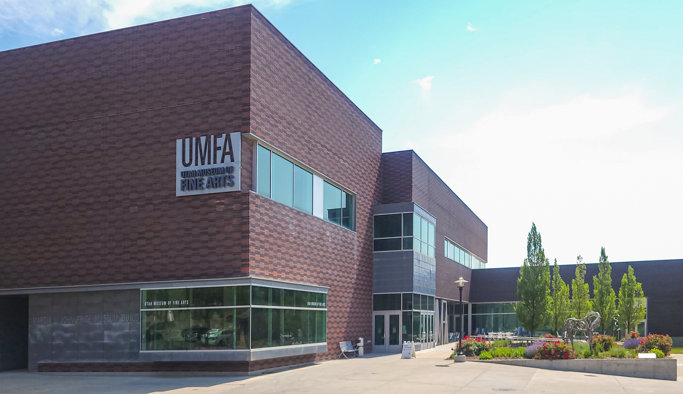A large, brown brick building has large glass windows. There's a sign that says UMFA in large metal letters on the side of the building. The sun is out and the sky is blue. There are green trees and plants to the side of the building.