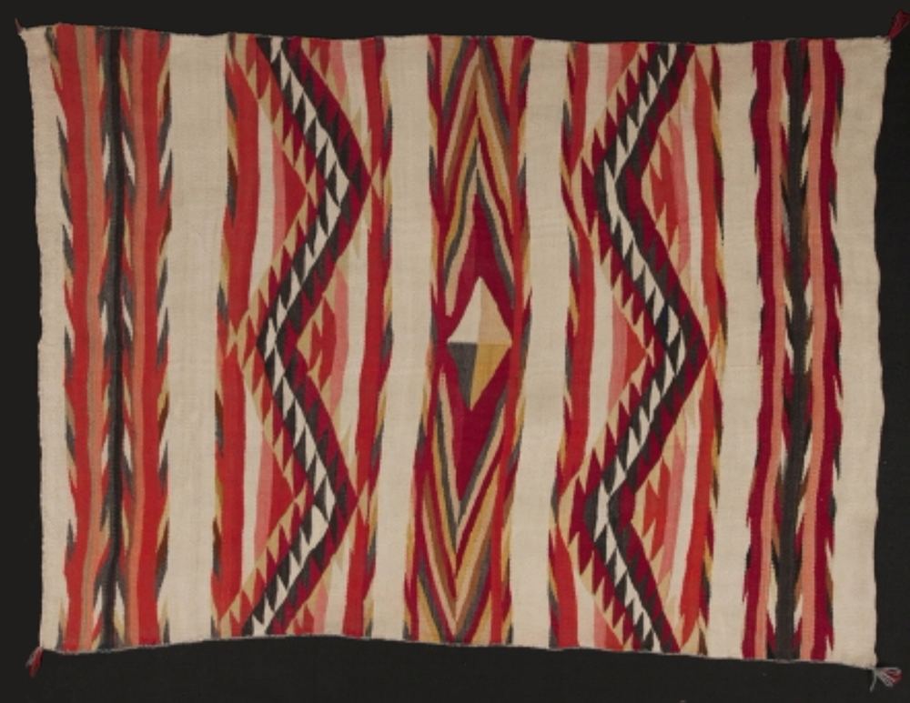 A woven rug with a bright geometric design. Red, pink, black, green and yellow lines zig zag across the rug in stripes. A diamond divided into quarters of different colors sits in the middle.