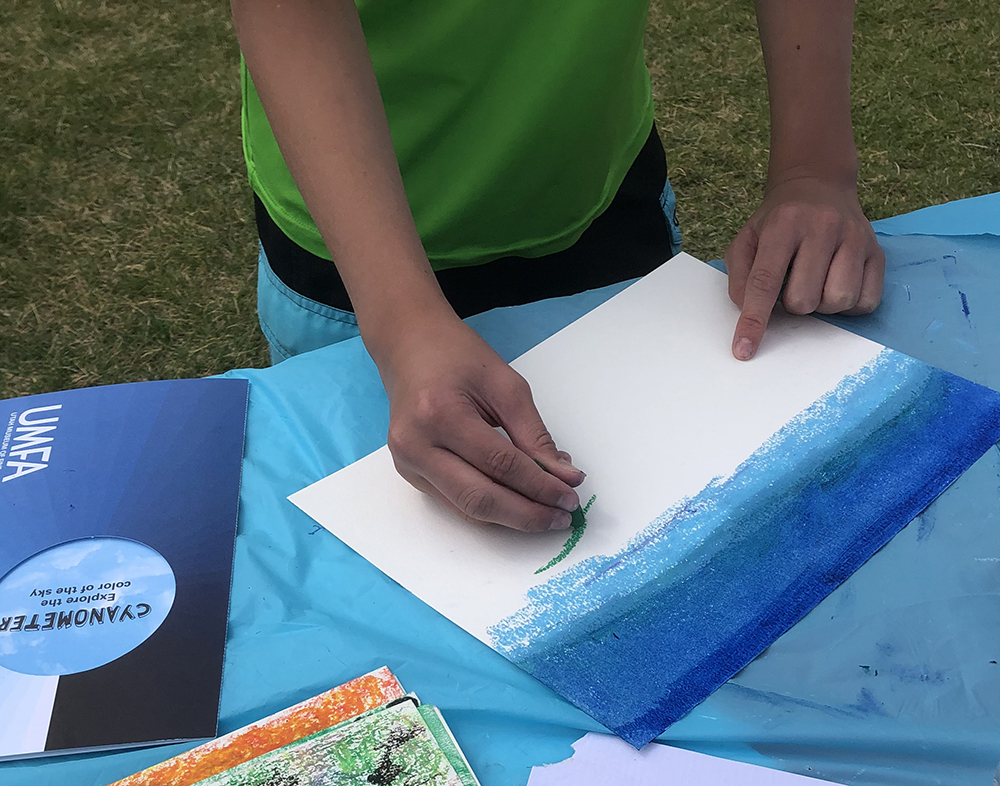 Child drawing a blue sky with crayons on a blue table only the child's hands and torso are in view, child is wearing a green shirt.