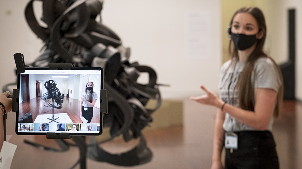 A UMFA educator standing in front of an iPad on a tripod next to Chakaia Booker's Discarded Memories sculpture  