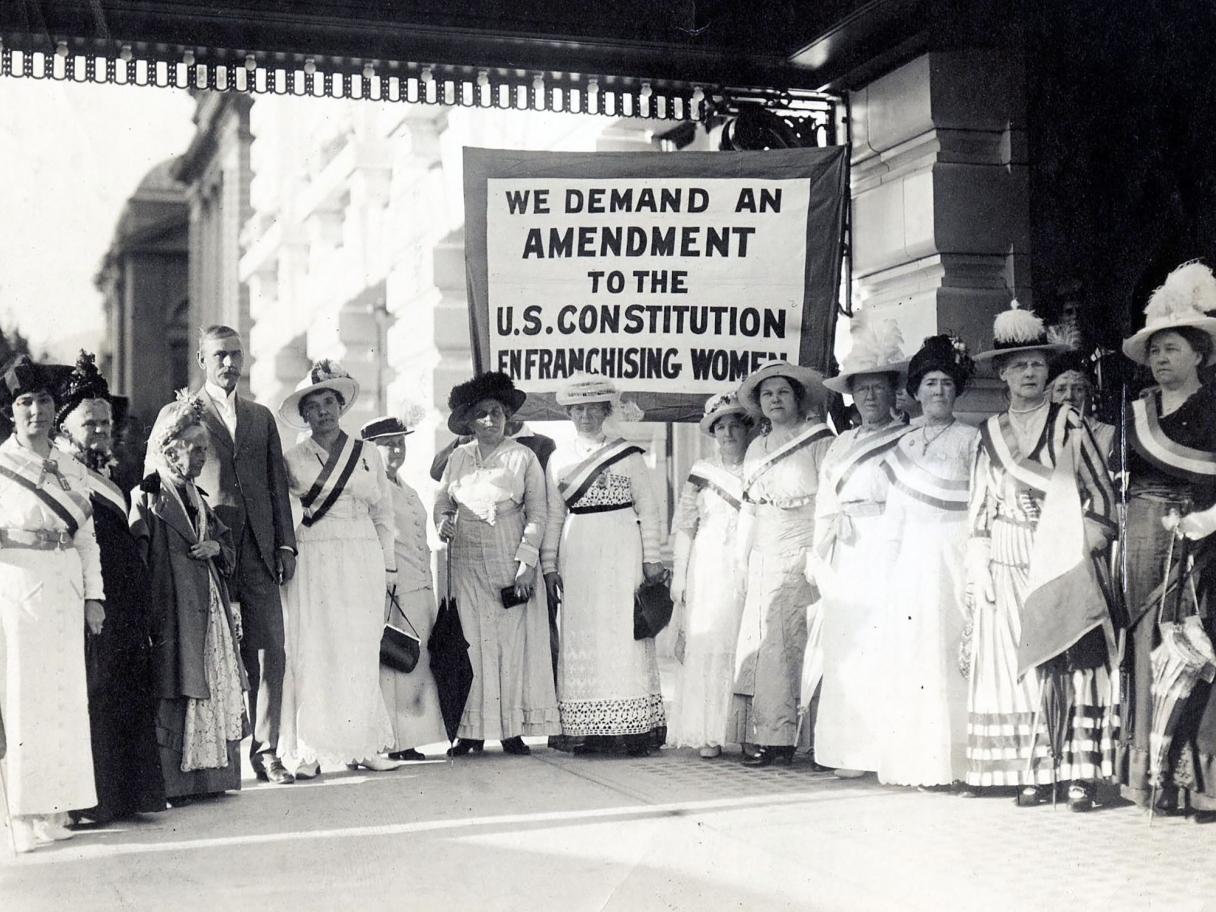 Susan B. Anthony stands with a group of  Women Suffrage Leaders in a group in front of Hotel Utah in 1915 behind the groups a banner reads We demand an amendment to the US Constitution Enfranchising Women. 