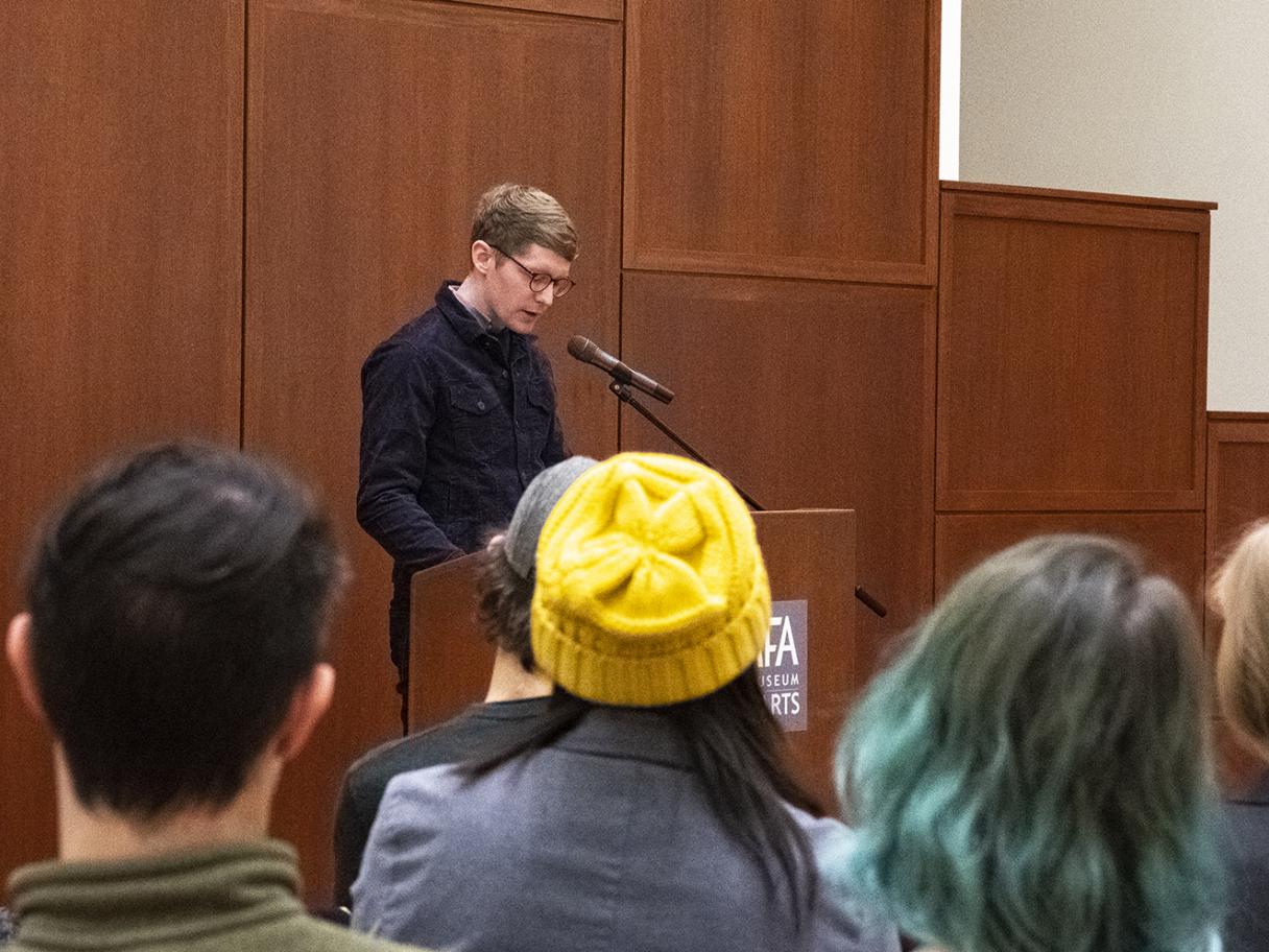 Working Dog February 2020 Working Dog reading, man a podium wearing a black shirt with a back of heads of crowd watching in UMFA Great Hall