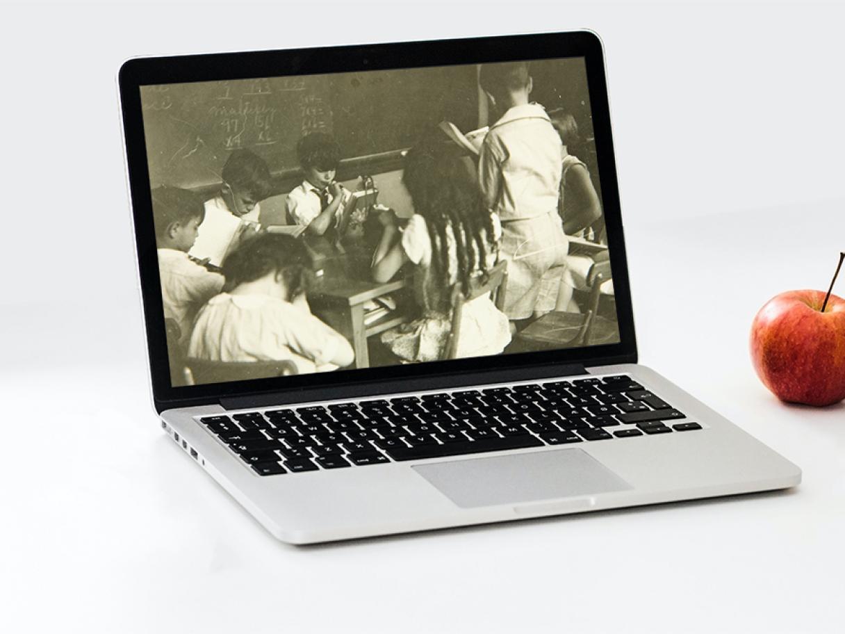 Laptop in an all white field, Olive Garrison photograph of children in school house on screen, and apple sits next to the computer. 