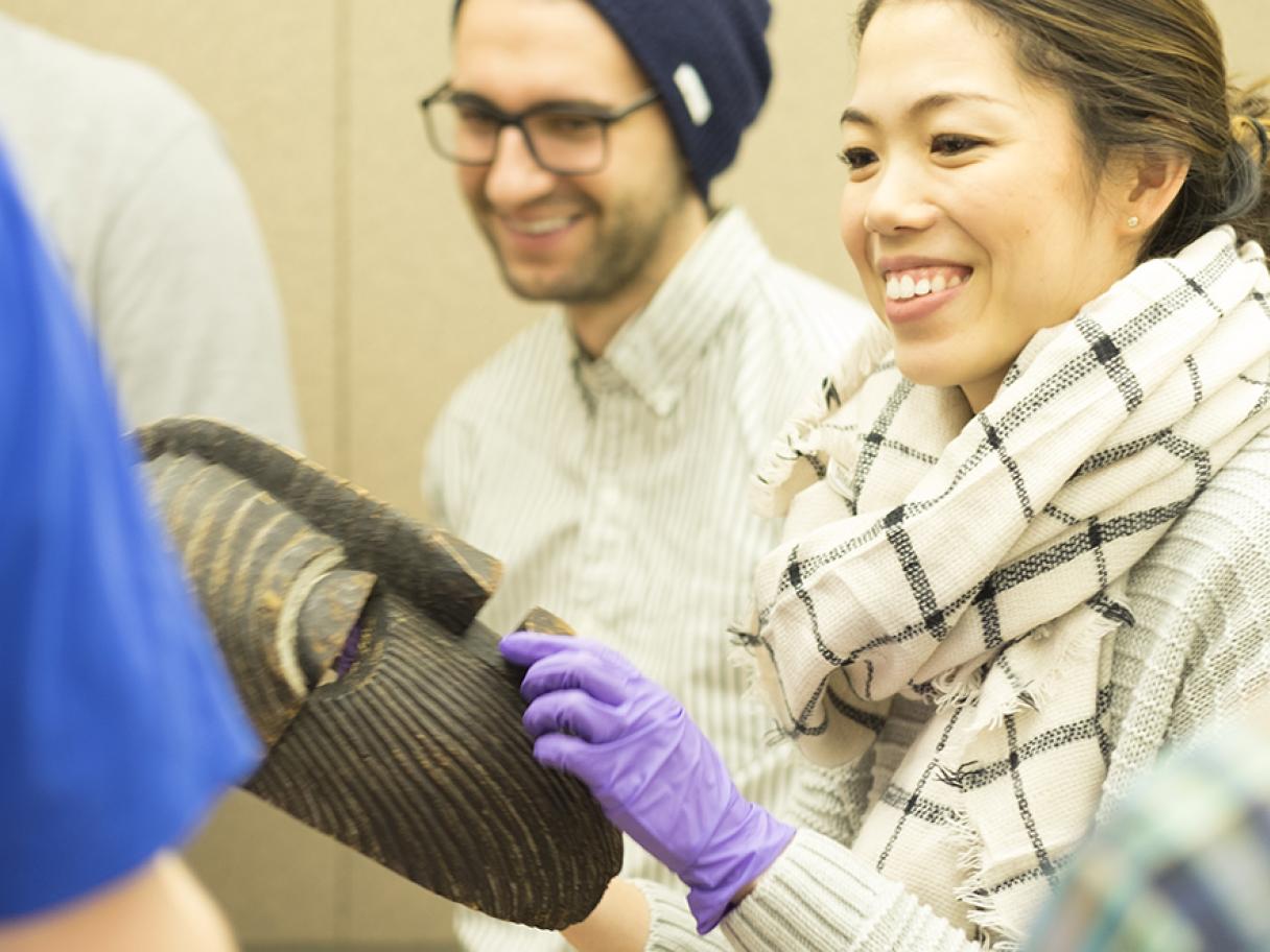 Female med student hold an African mask durring a visual thinking strategies class she wears purple nitrile gloves, white scarf with black squares, her hair is pulled back in a ponytail.