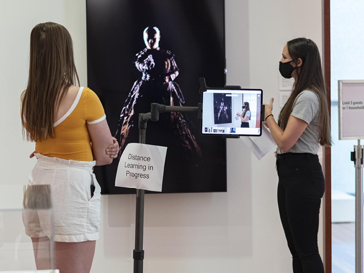 two UMFA educators stand in front of Yuki Kihara's Shiva in Motion, an iPad on a stand films on of the educators, she wears a grey shirt and black face mask