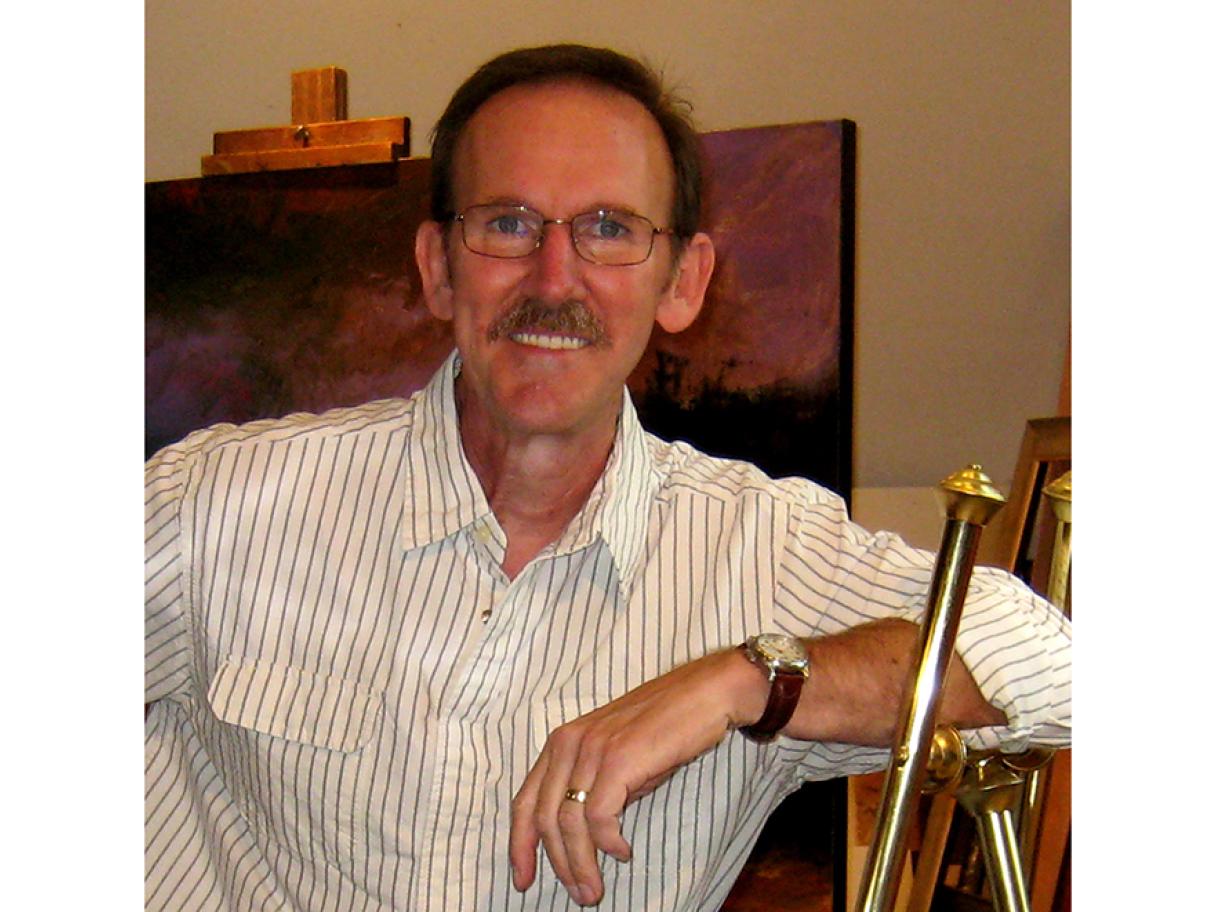 Don Prys Don Prys stands with arm leaning on easel he wears a white button-up shirt with black pin stripes a painting is behind him