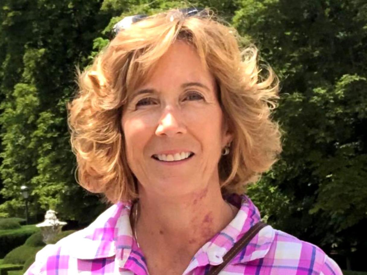 Lori Scharf a woman with medium length light brown hair wearing a pink and purple plaid shirt standing in front of a green hedge