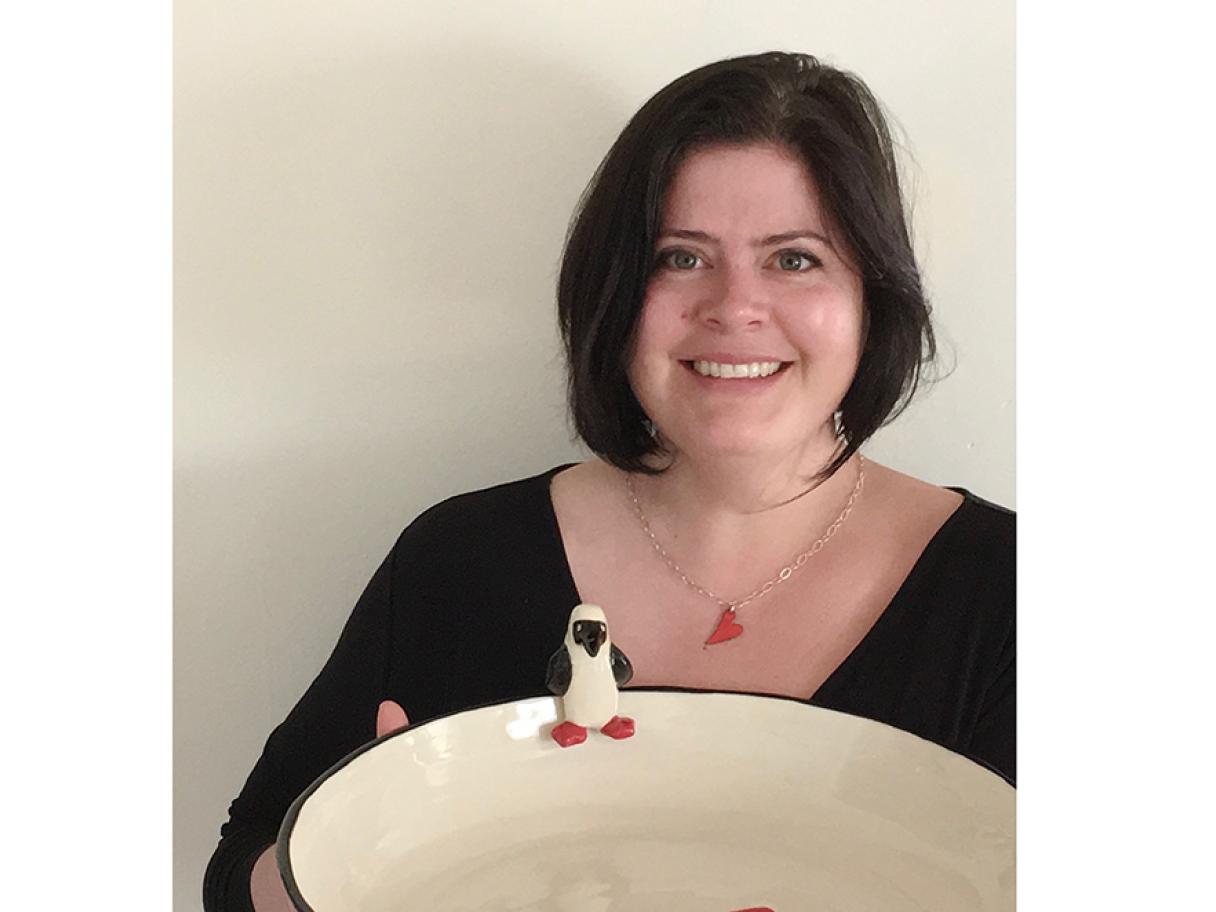 Mira Loyborg has dark brown hair in a bob cut wears black shirt with v-neck holding large white bowl with ceramic penguin on the rim all in front of a white wall 