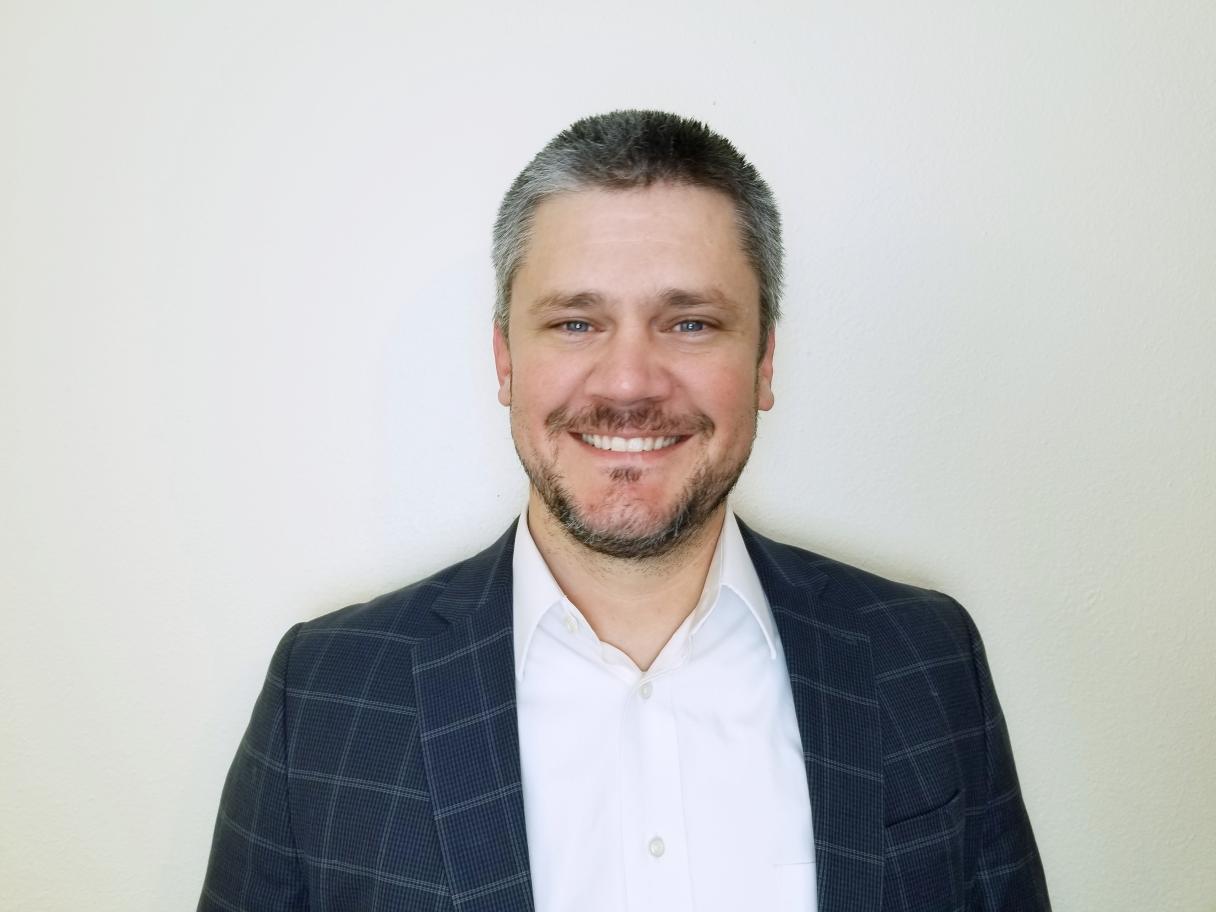 Ron Fehr headshot smiling at camers wearing white shirt and blue plaid jacket. He has short graying hair and short beard