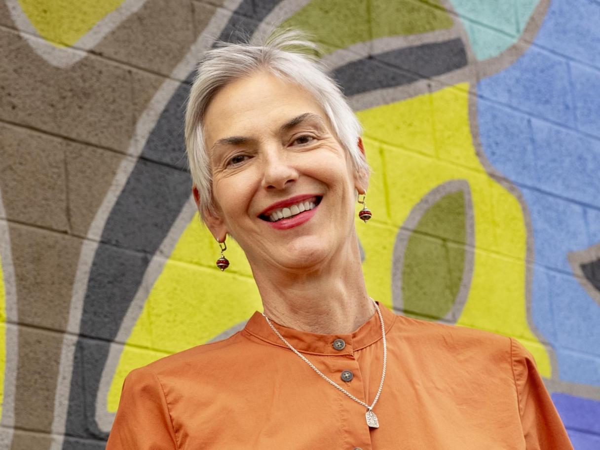 Traci Overy Covey a woman with short gray hair wearing an orange shirt standing in front of a mural