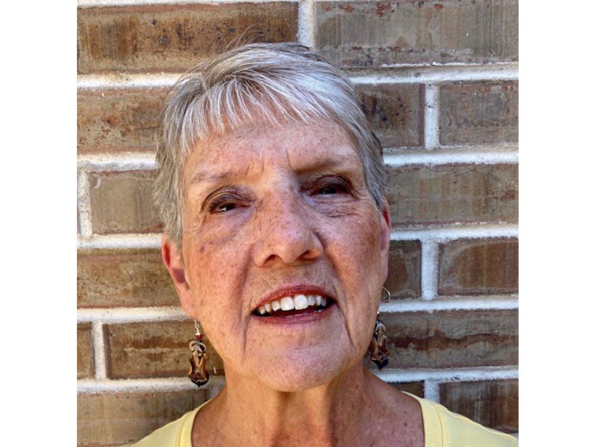 an older woman with short gray hair stands against a brick wall she is wearing a light yellow shirt