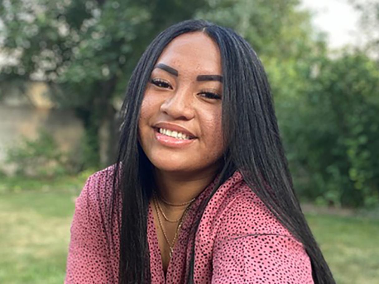 Monique Ho Ching A young woman with long black hair wearing a dark pink blouse sitting outside