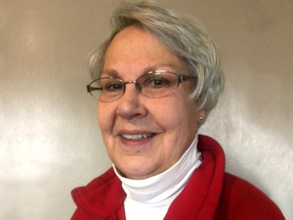 Rita Egbert an older woman with short gray hair and oval framed glasses, wearing  a white turtleneck and red sweater  