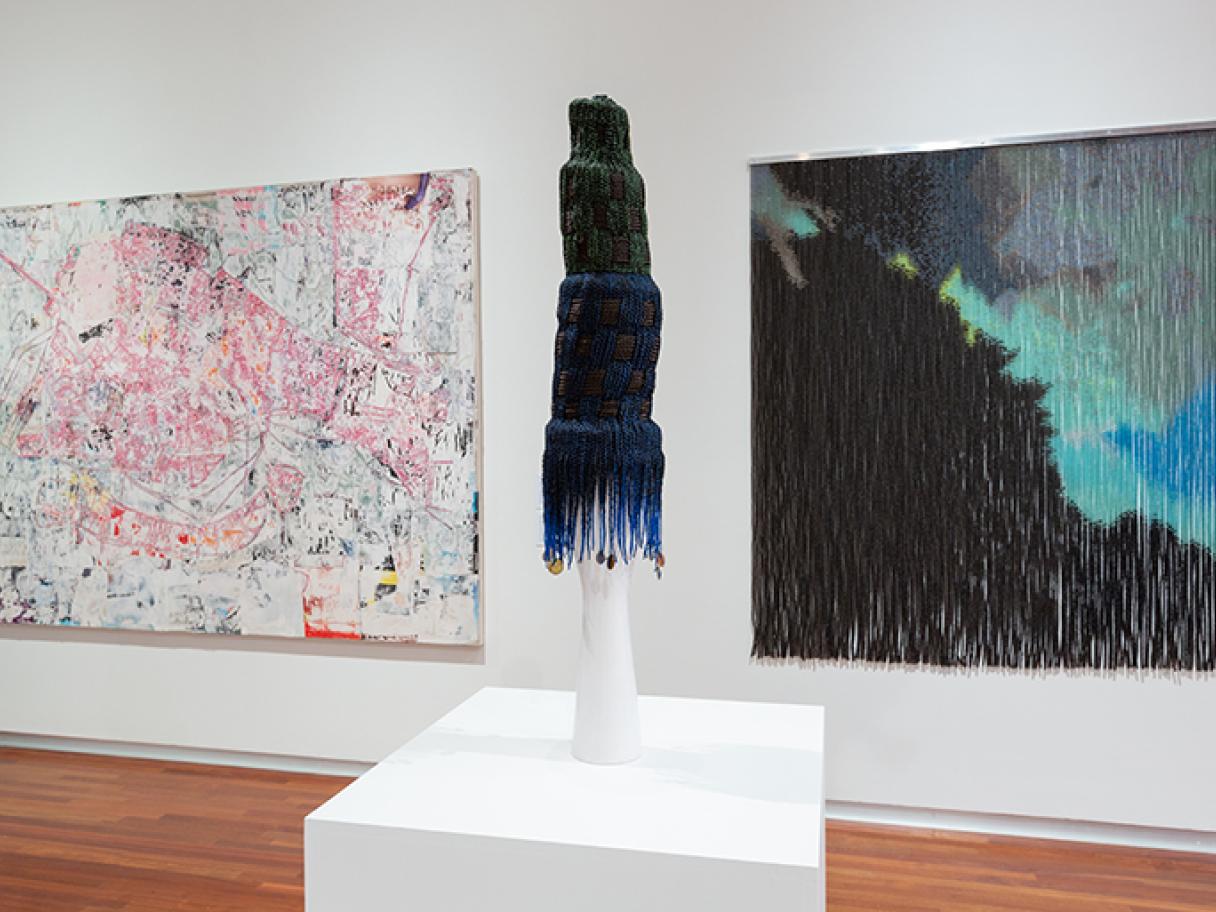 A sculpture called Lipstick Building made of braided synthetic hair sits on a white pedestal two abstract pieces of art hang on the wall behind  