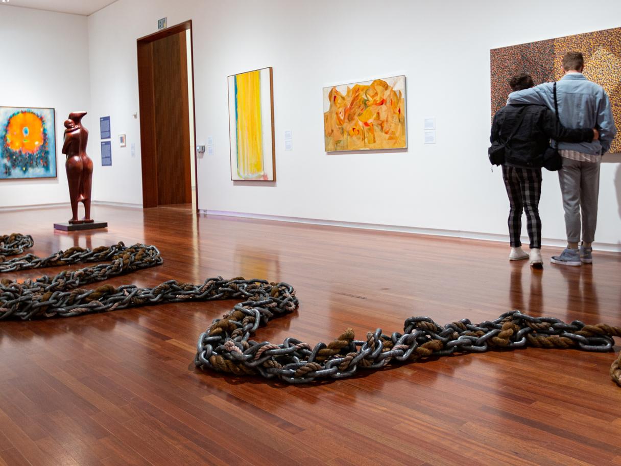 Black Refractions gallery a sculpture of chains and ropes snakes across the floor a coupld stands with arms wrapped around each other looking an abstract painting 