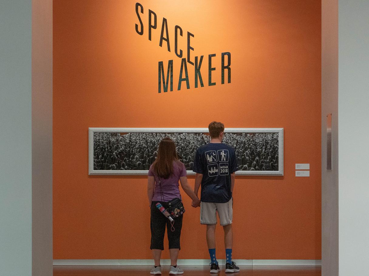 Space Maker title wall with couple a couple holding hands looking at a black and white, narrow, horizontal photo on an orange wall with Space Maker title on the wall above