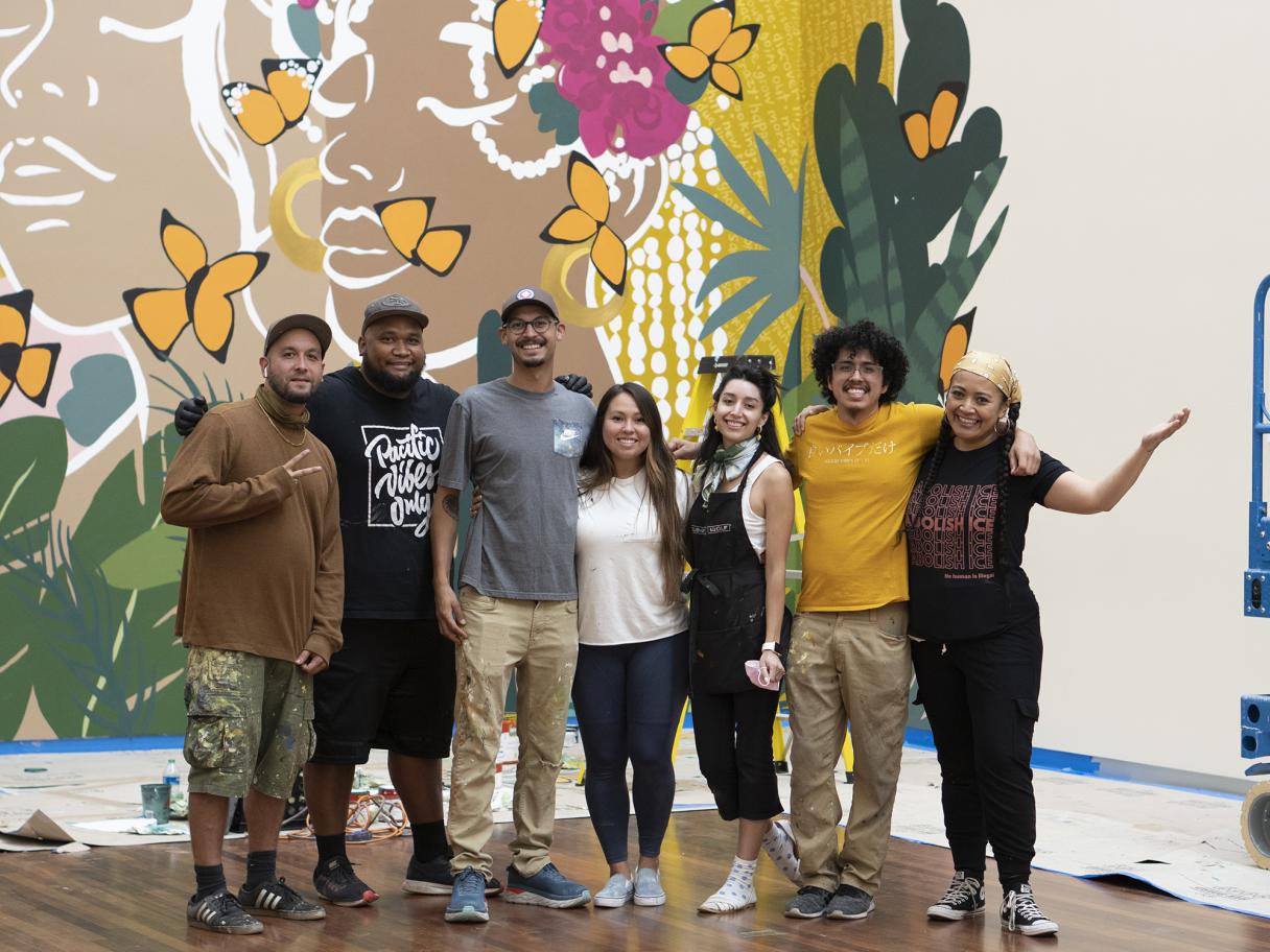 The seven muralists of 2020: From here on out posing infront of a mural in the UMFA Great Hall 