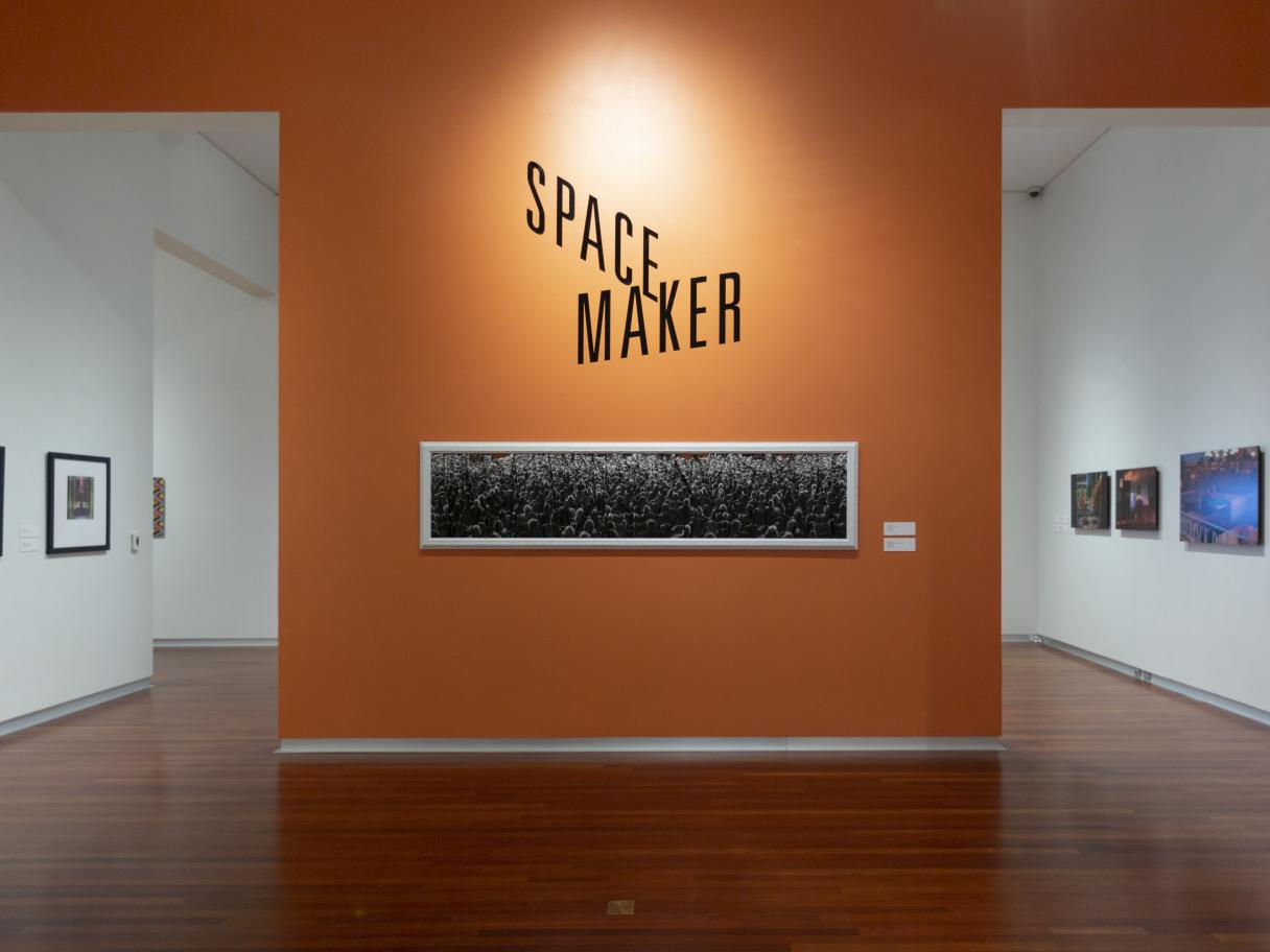 Title wall in the UMFA gallery with Space Maker in larger black text on an orange wall, above a long horizontal black and white photo.