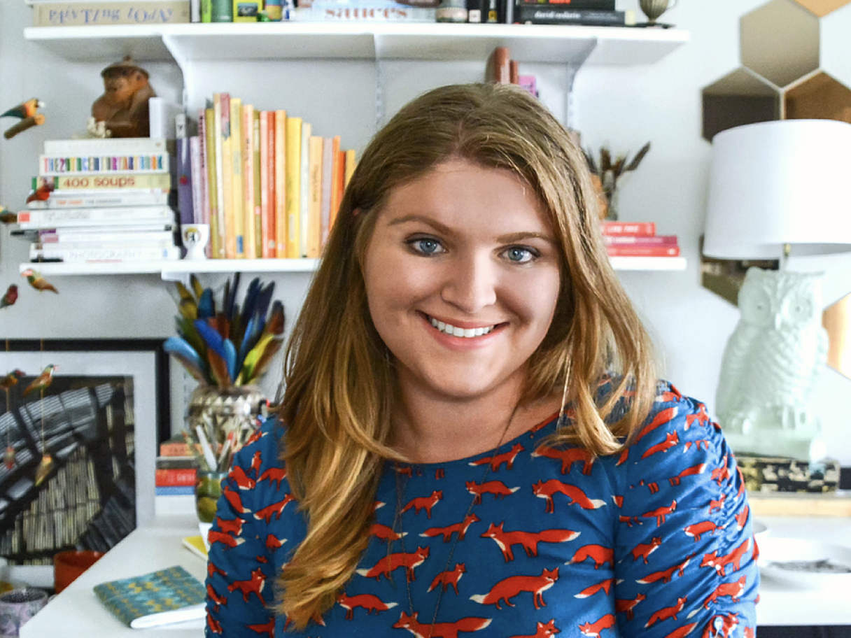 Rachel Kozlowski Rachel Kozlowski sits infrom of a bookshelf wearing a blue shirt with an all-over print of red foxes she has long dark blond hair