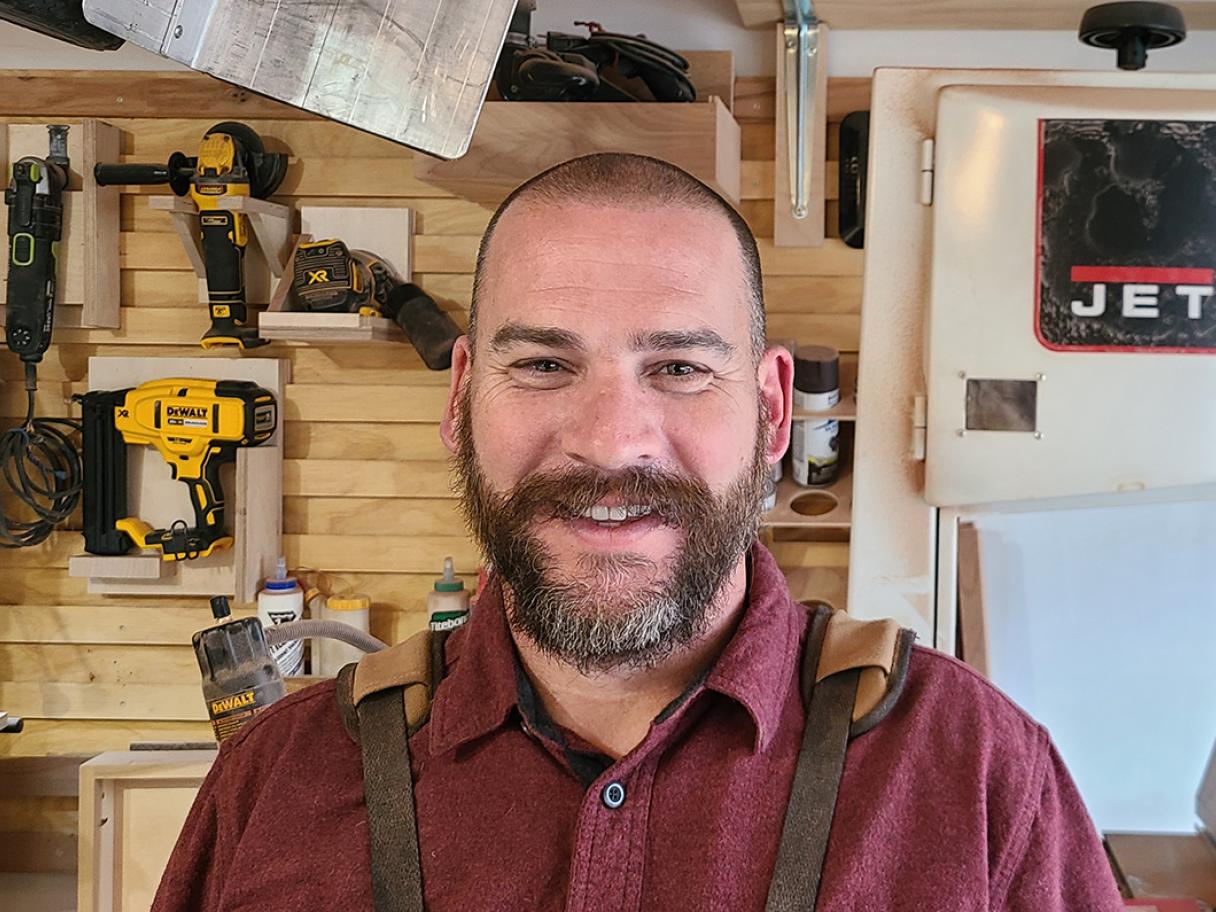 Tyler Smith Tyler Smith stands in his wood shop he's wearing a red button up shirt and brown suspenders.