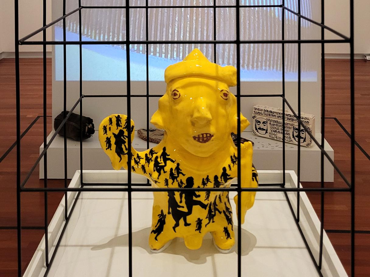 a replica of an ancient mesoamerican sculpture of a child glazed bright yellow with a pattern of silhouetted children across the body, the sculoture sits in a black cage 