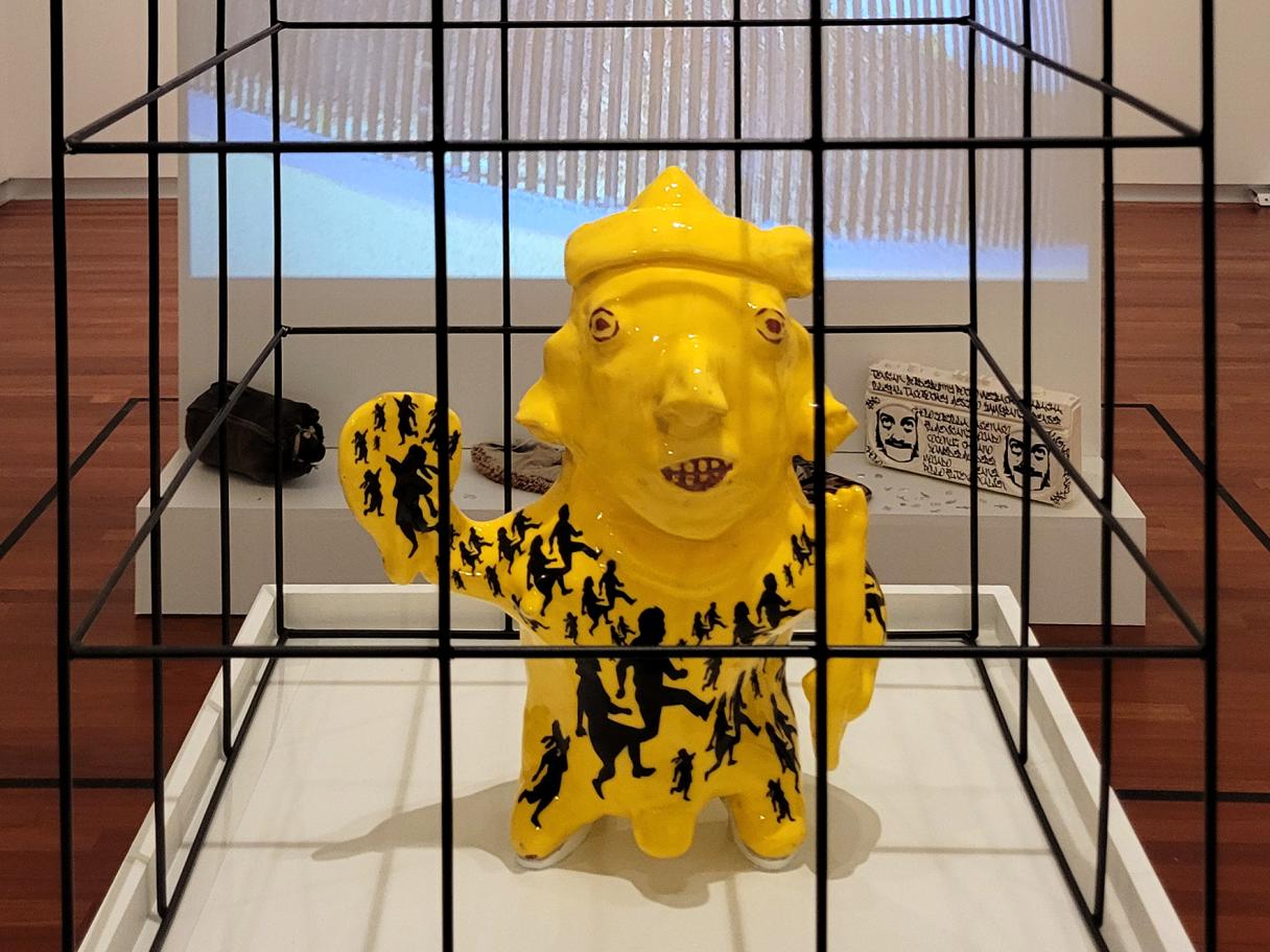 Salt 15 hero 1 a replica of an ancient mesoamerican sculpture of a child glazed bright yellow with a pattern of silhouetted children across the body, the sculoture sits in a black cage